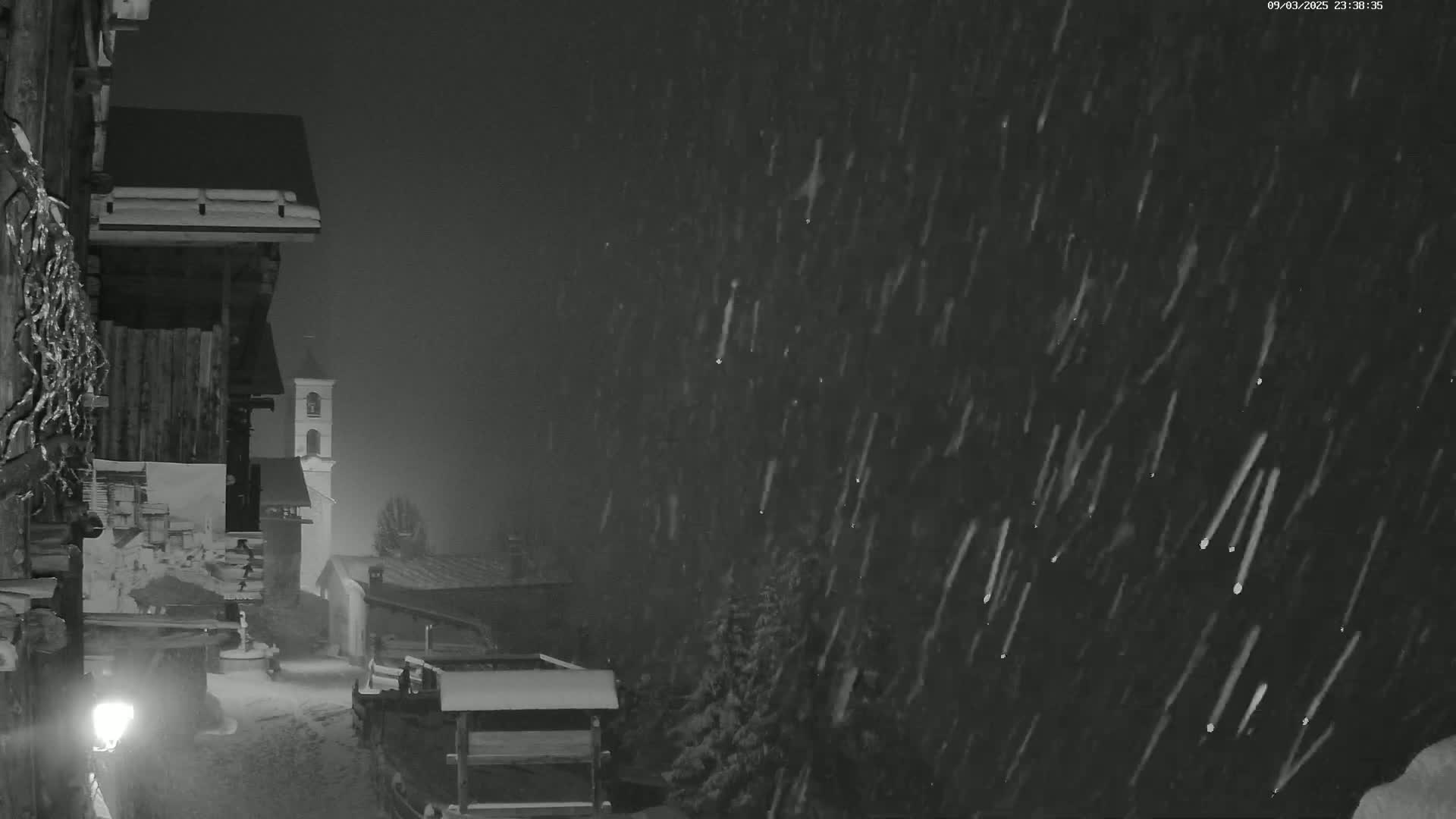 A nighttime scene of snow falling heavily on a village with old wooden buildings, including a church steeple, partially visible through the snow.