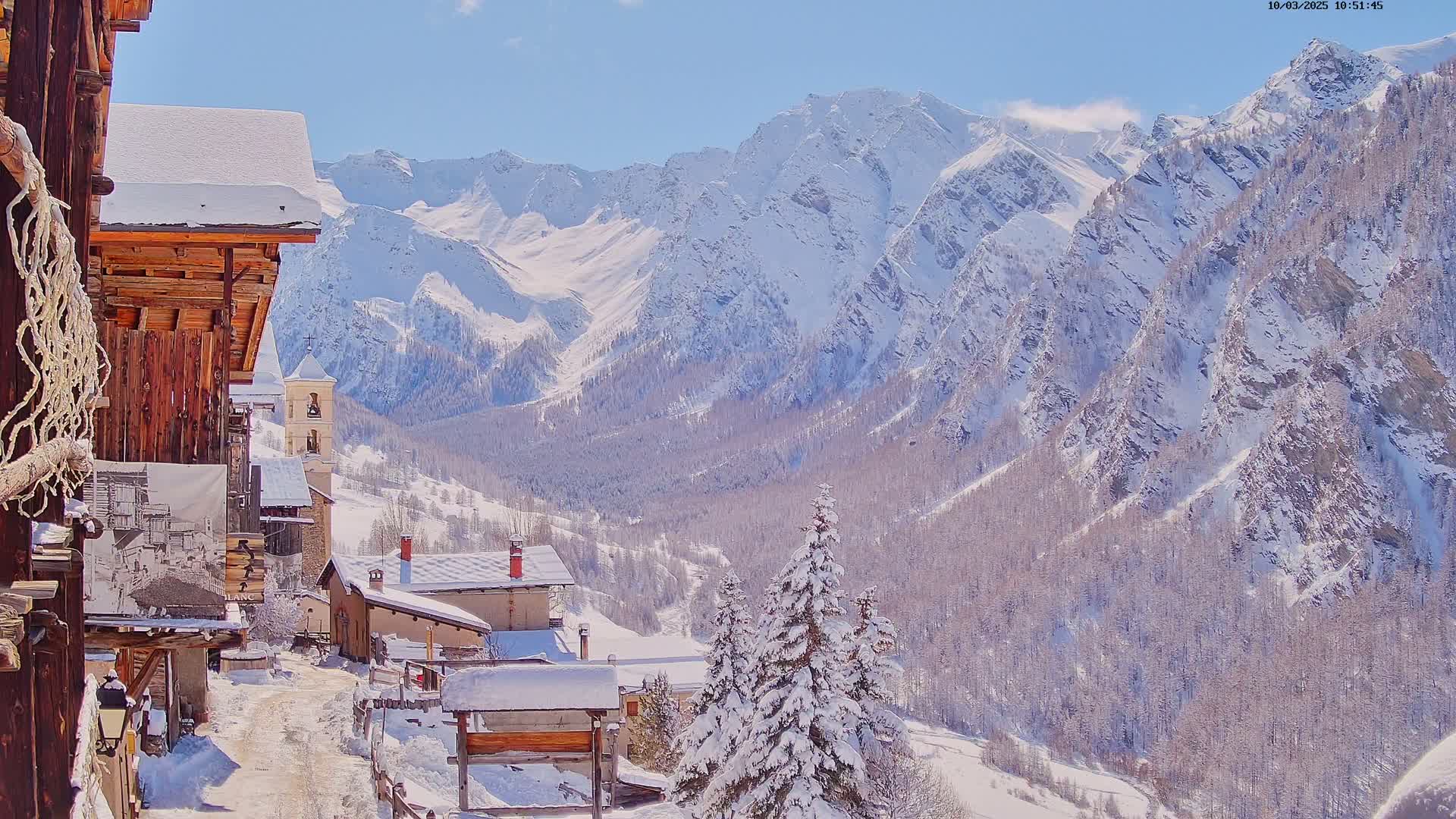 A snow-covered village nestled in a valley, overlooked by a towering, snow-capped mountain range under a bright, sunny sky.