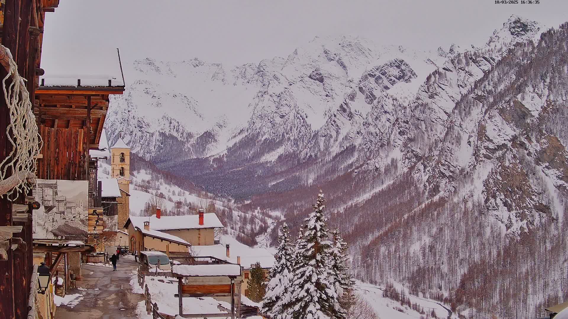 A snow-covered village nestled in a valley, overlooked by towering, snow-capped mountains under an overcast sky.
