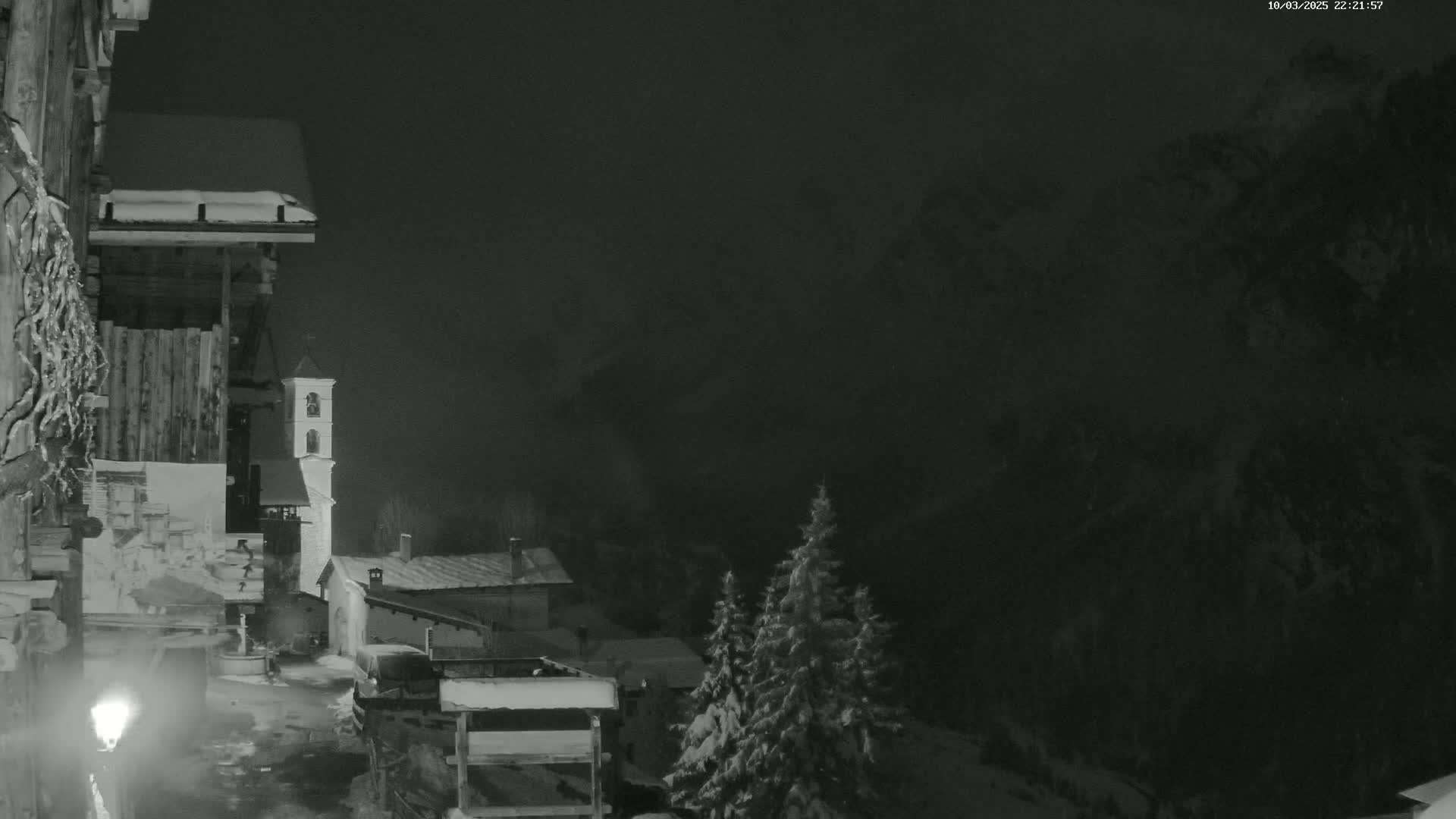 A snow-covered village at night is illuminated by a streetlamp, with a church steeple visible in the distance and dark, snow-covered mountains in the background.