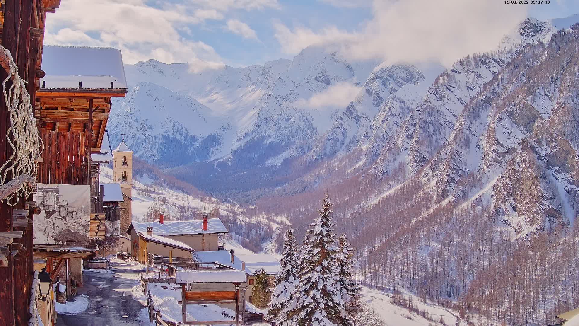 A snow-covered village nestled in a valley, overlooked by a towering, snow-capped mountain range under a partly cloudy sky.