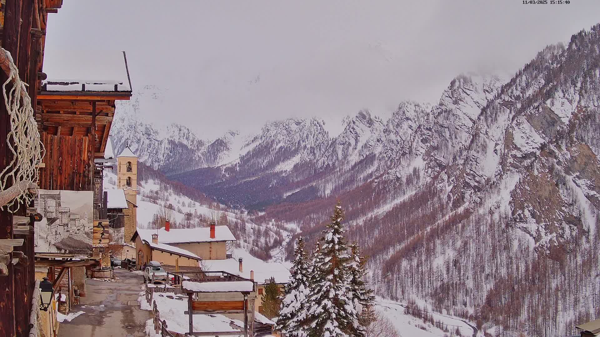 A snow-covered village nestled in a valley between towering, snow-capped mountains under an overcast sky.