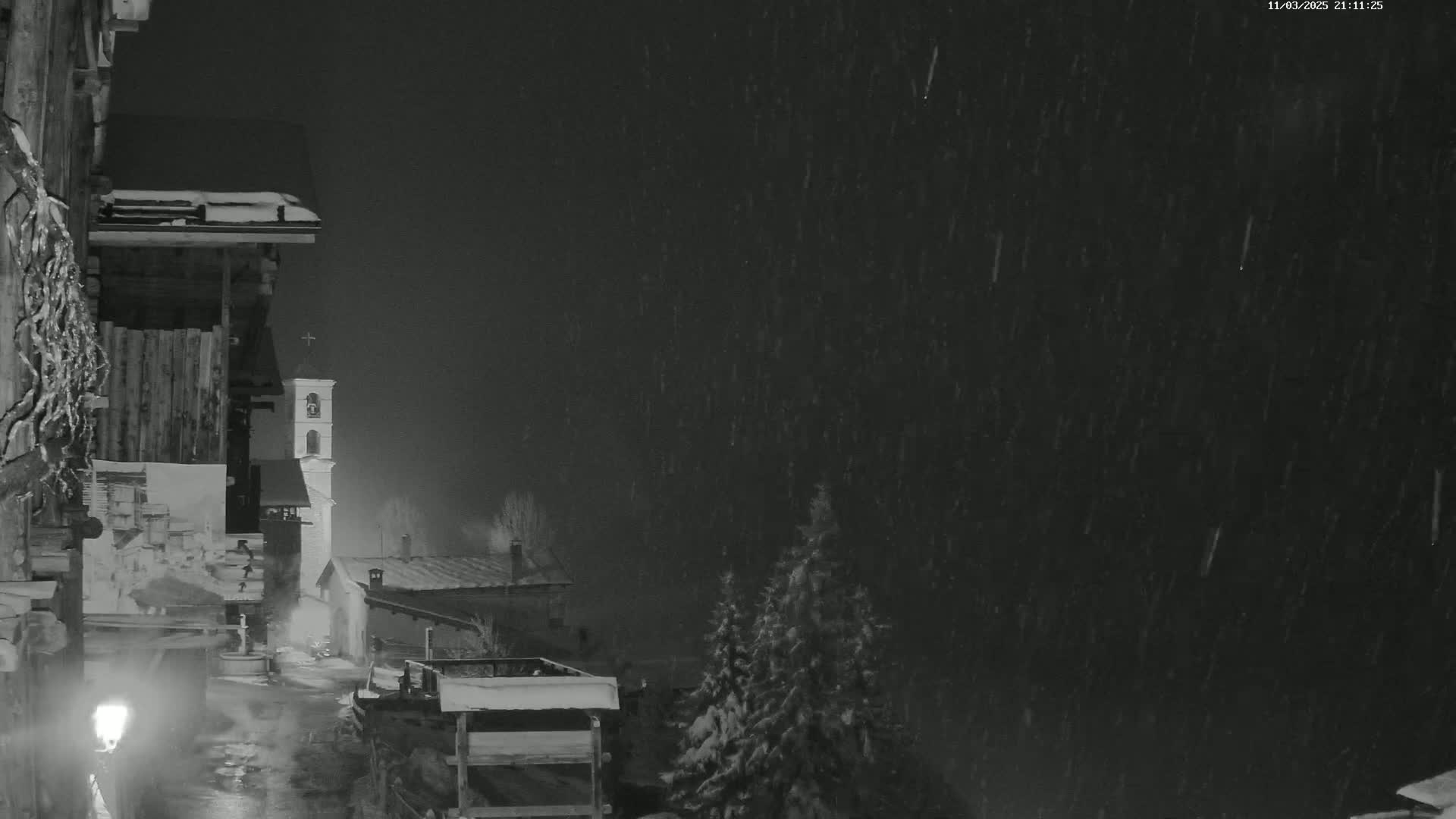 A nighttime scene of snow falling on a village with a church steeple visible in the distance, illuminated by a single lamppost.