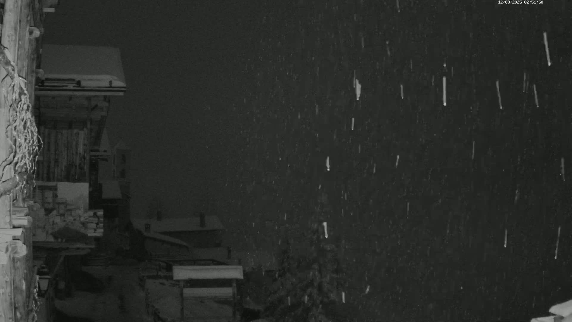 A nighttime scene of snow falling heavily over a snow-covered building and surrounding landscape.