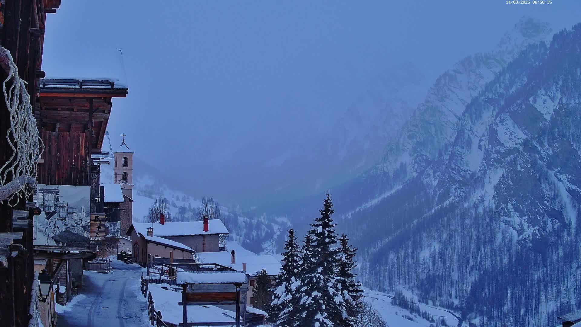 A snow-covered village nestled in a valley, overlooked by hazy, snow-capped mountains.