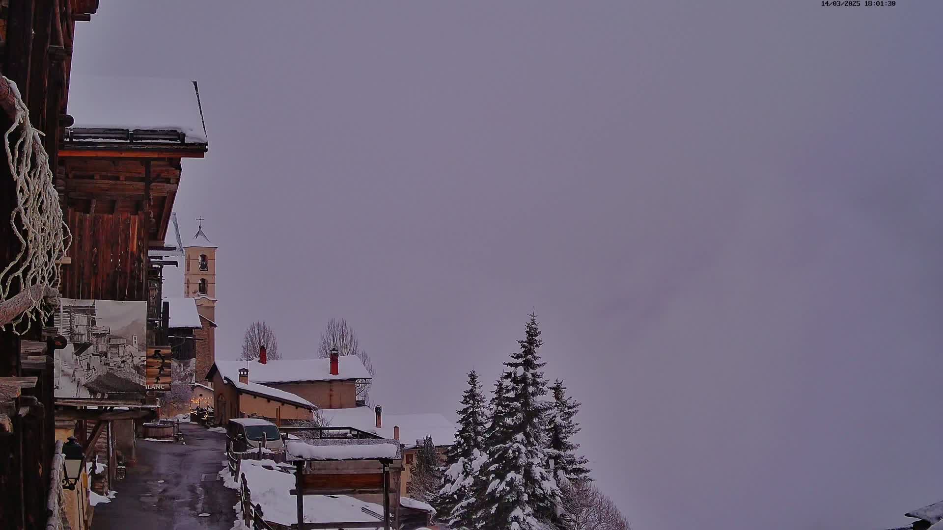 A snow-covered village street winds toward a church under a cloudy, overcast sky.