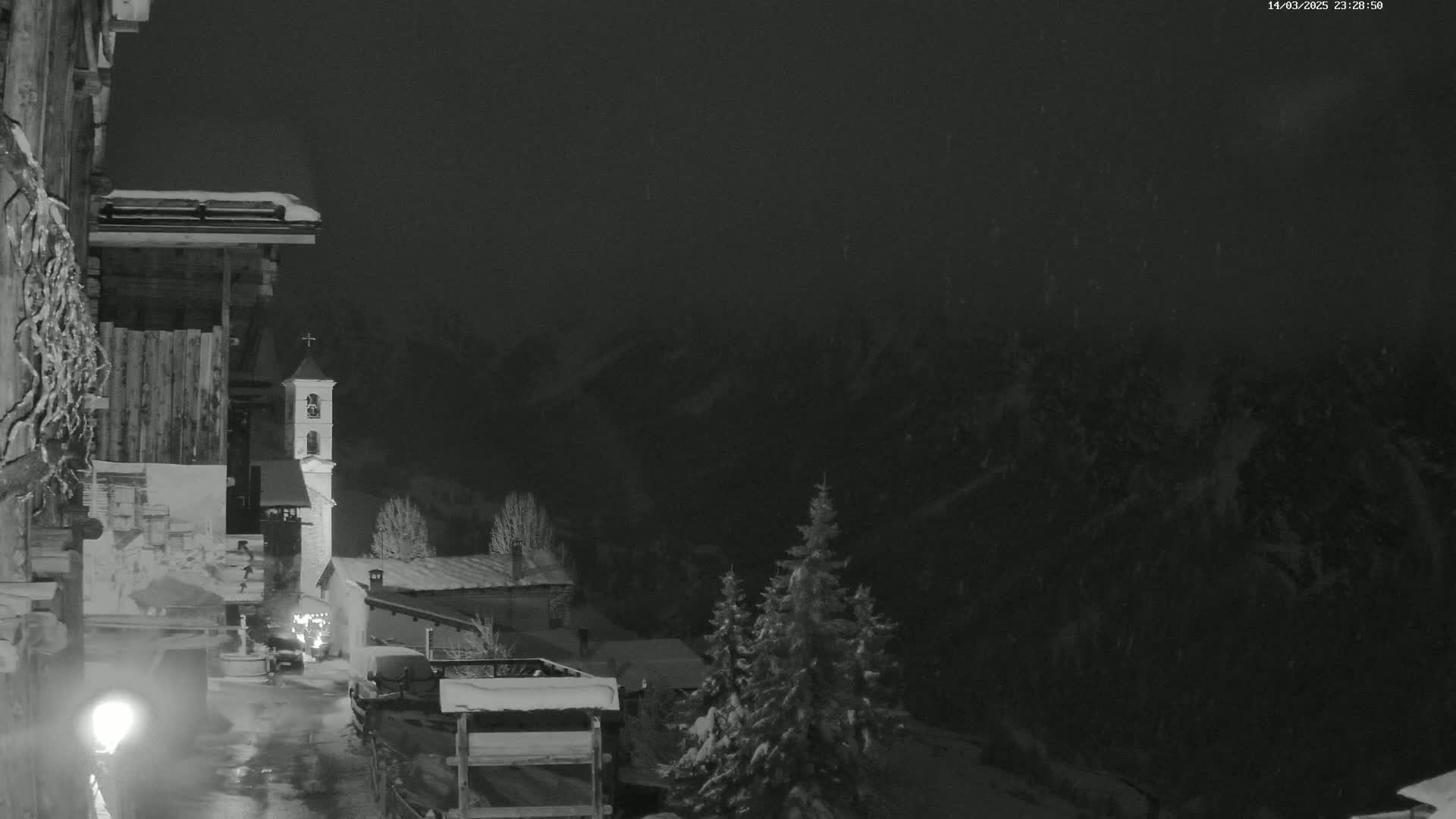 A snow-covered village at night, with a church steeple visible, is seen under falling snow.