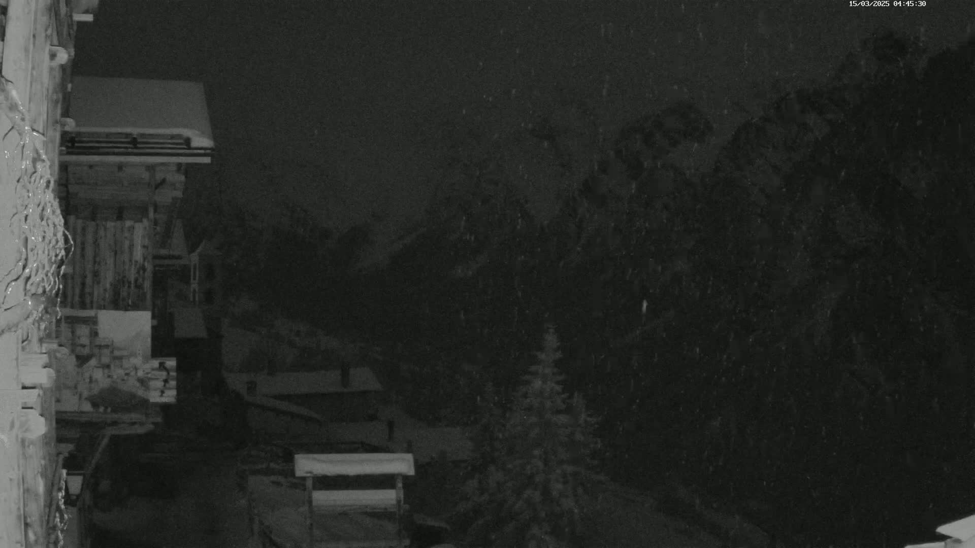 A nighttime view of snow falling on a snow-covered mountainous landscape, with a wooden building in the foreground.