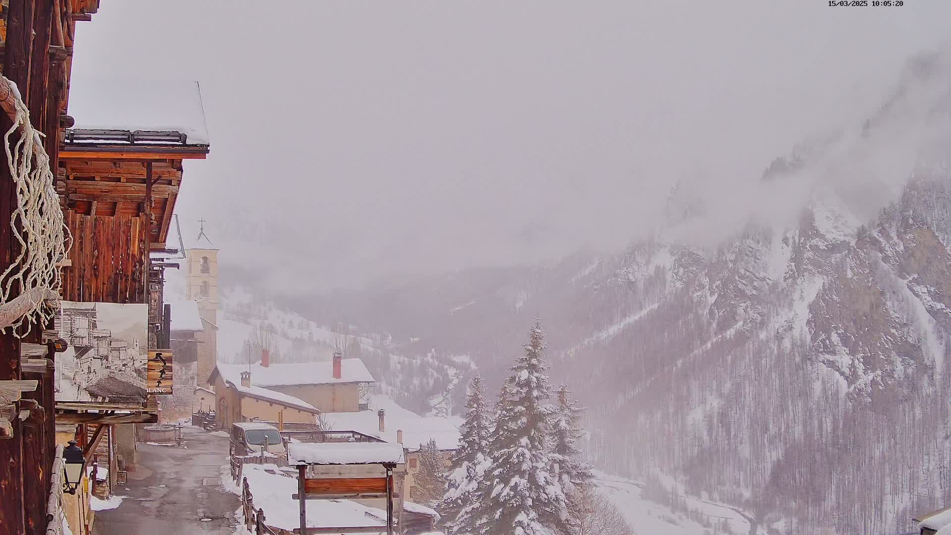 A snow-covered village nestled in a valley, viewed from a wooden building, with a foggy, snowy mountain range in the background.