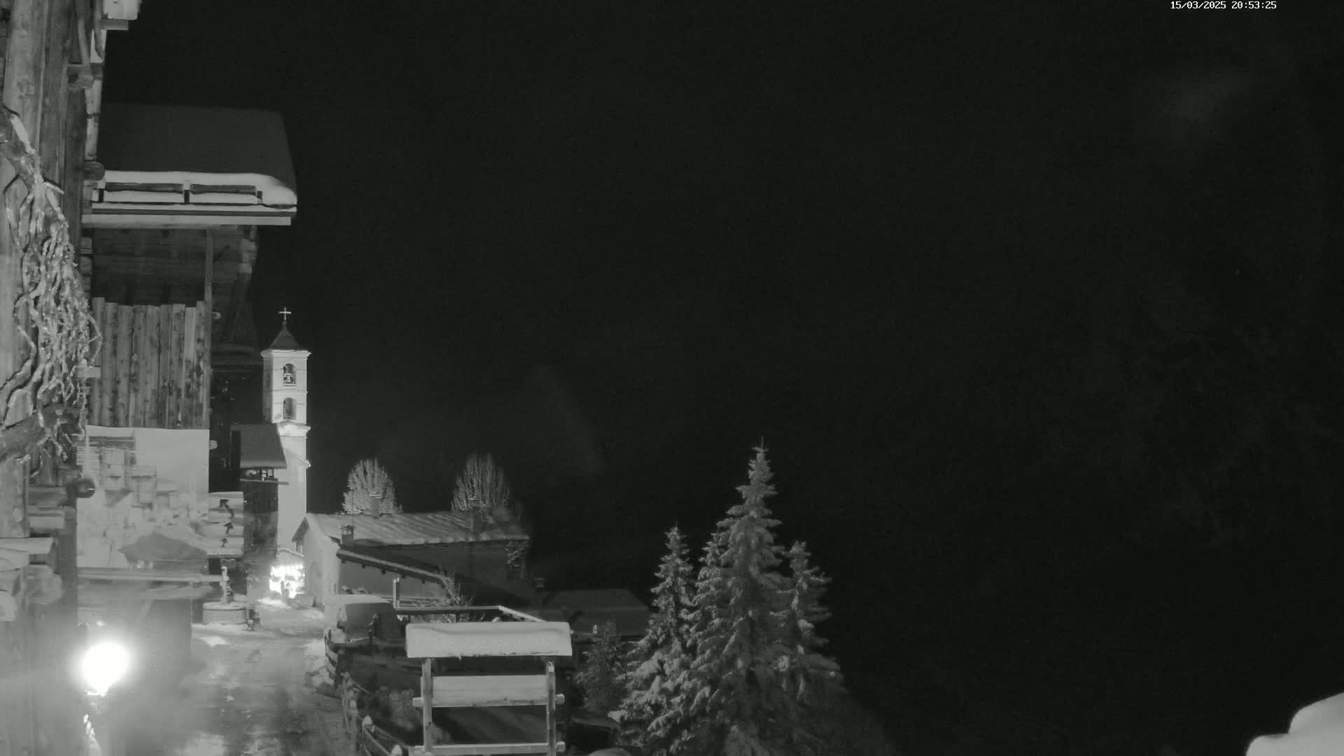 A snow-covered village at night, illuminated by a lamppost, features a church bell tower and several evergreens dusted with snow under a dark sky.