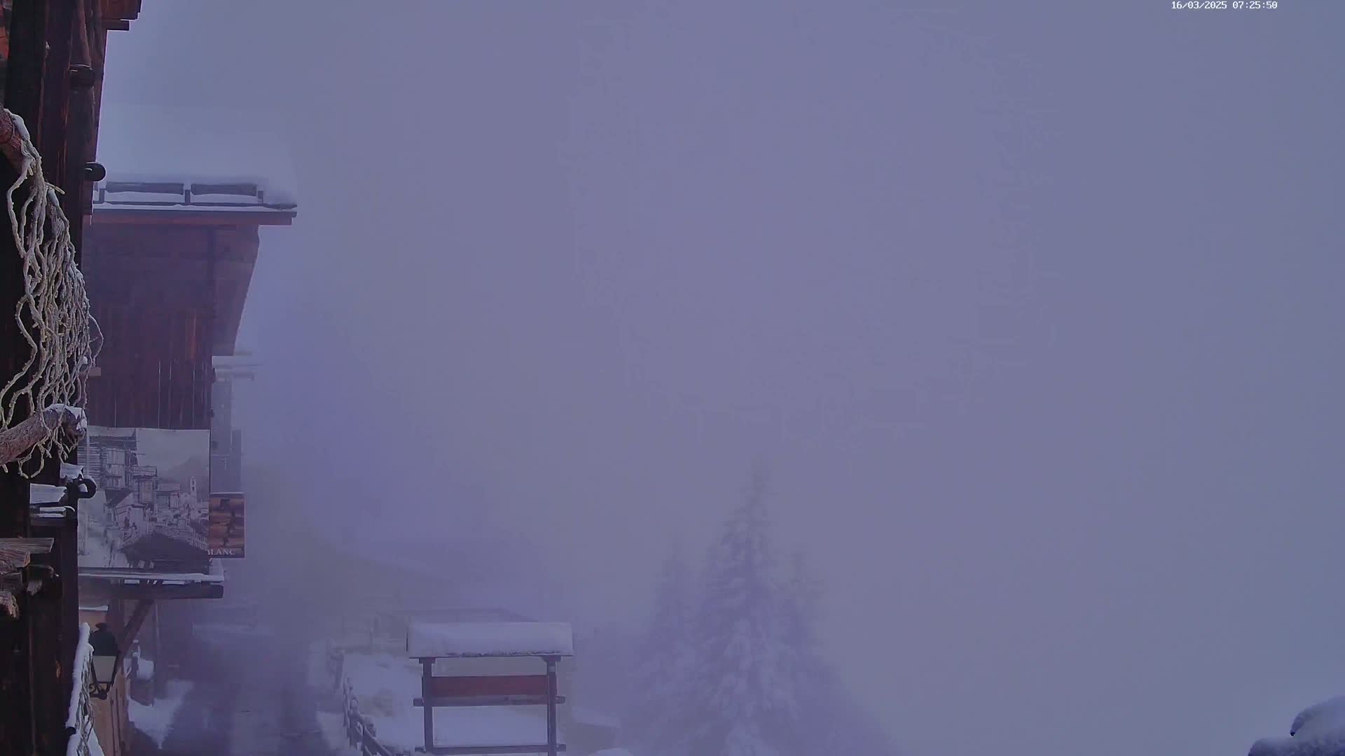 A snow-covered, wooden building is partially visible through a thick fog, with snow-dusted trees and other structures barely discernible in the distance.