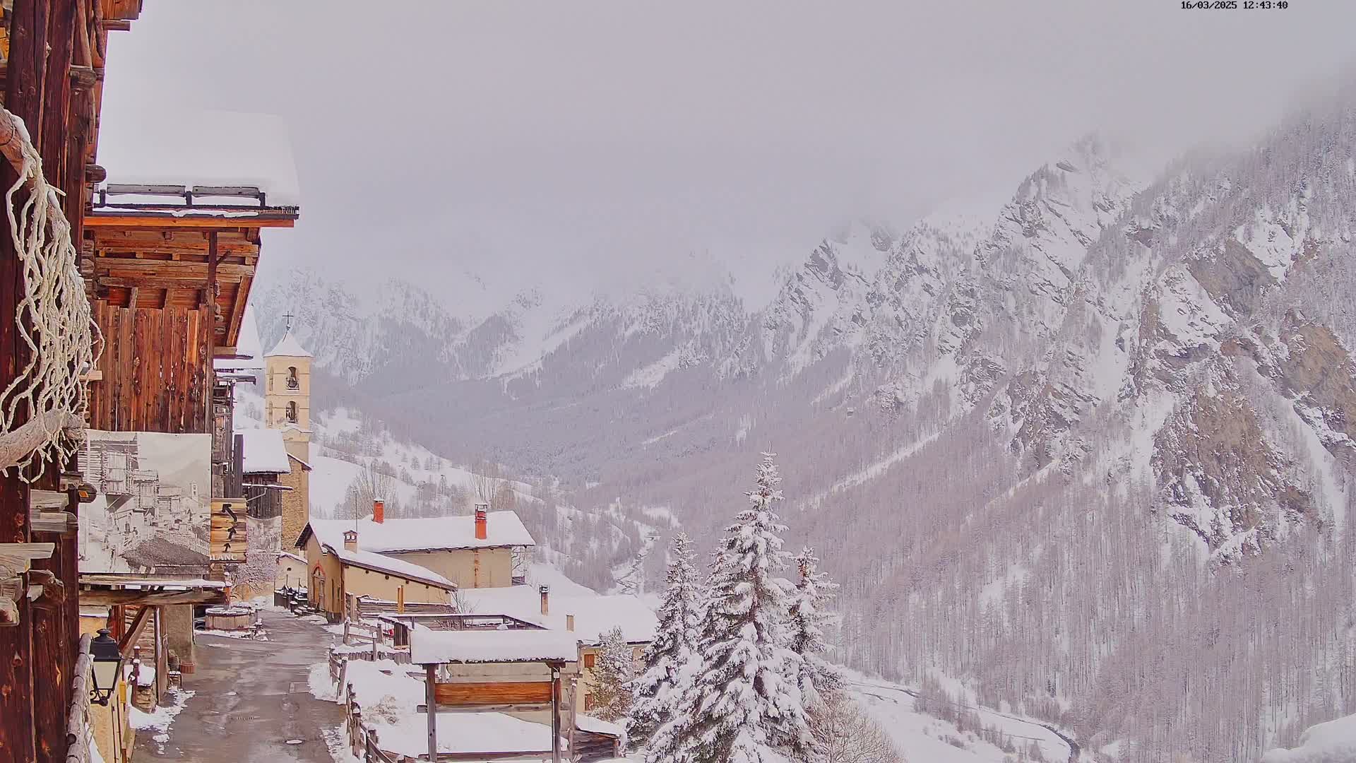 A snow-covered village nestled in a valley, overlooked by snow-capped mountains under an overcast sky.