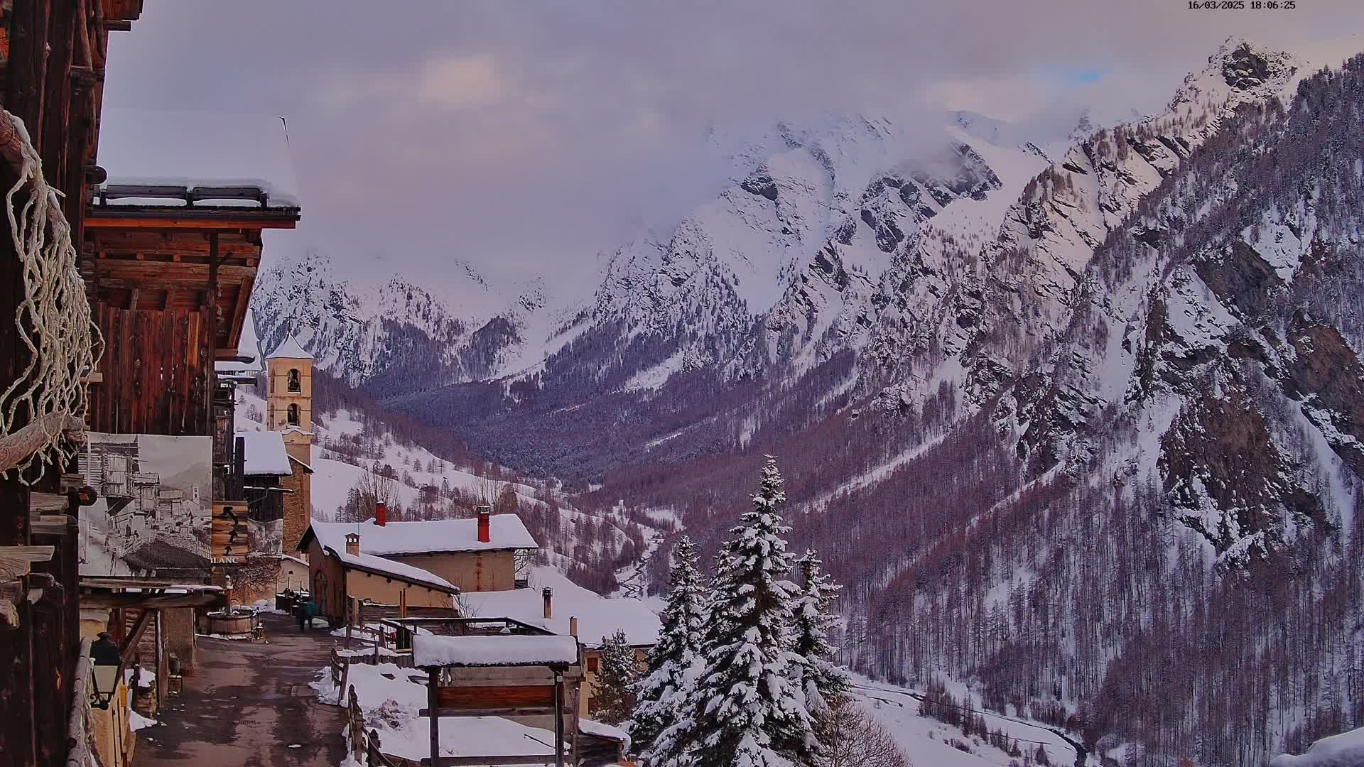 A snow-covered village nestled in a valley, overlooked by towering, snow-capped mountains under a cloudy sky.
