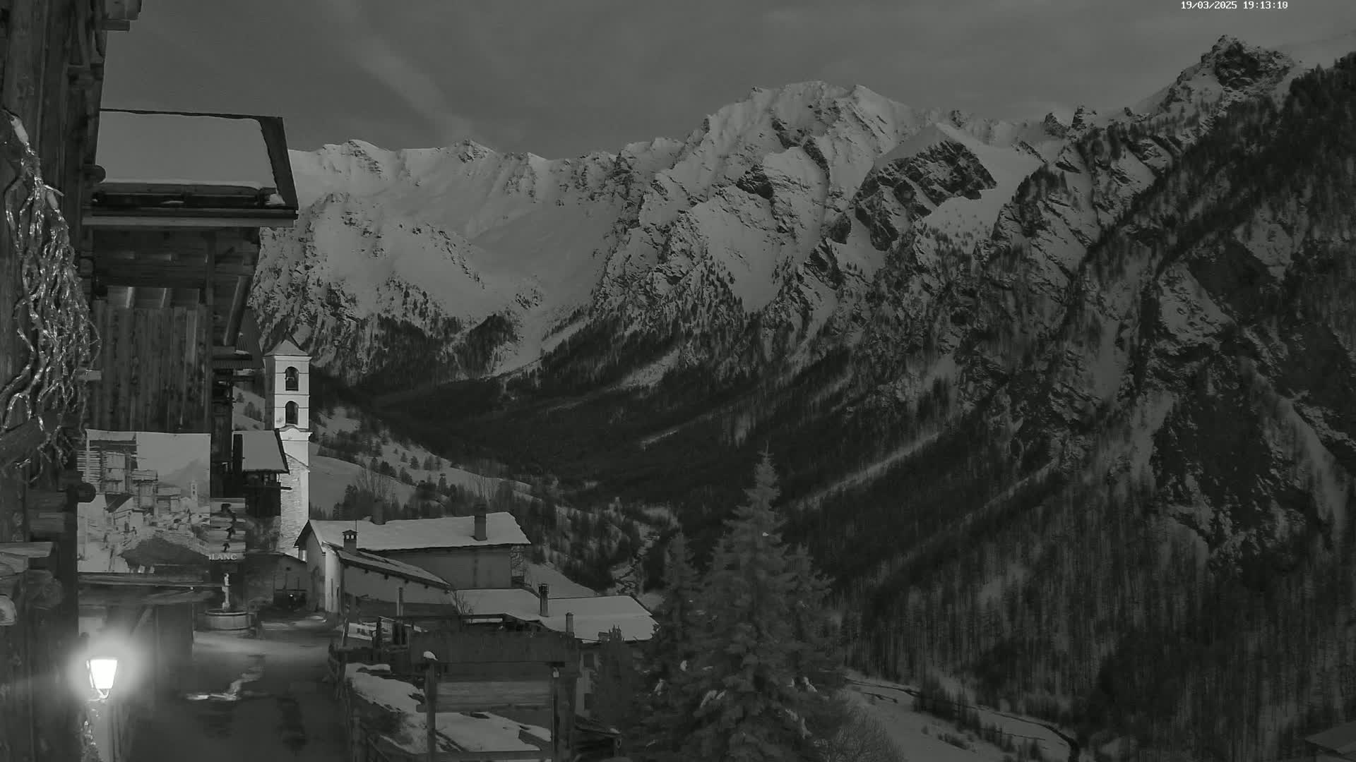 A snow-covered mountain range is seen from a village at night, lit by a single lamppost.