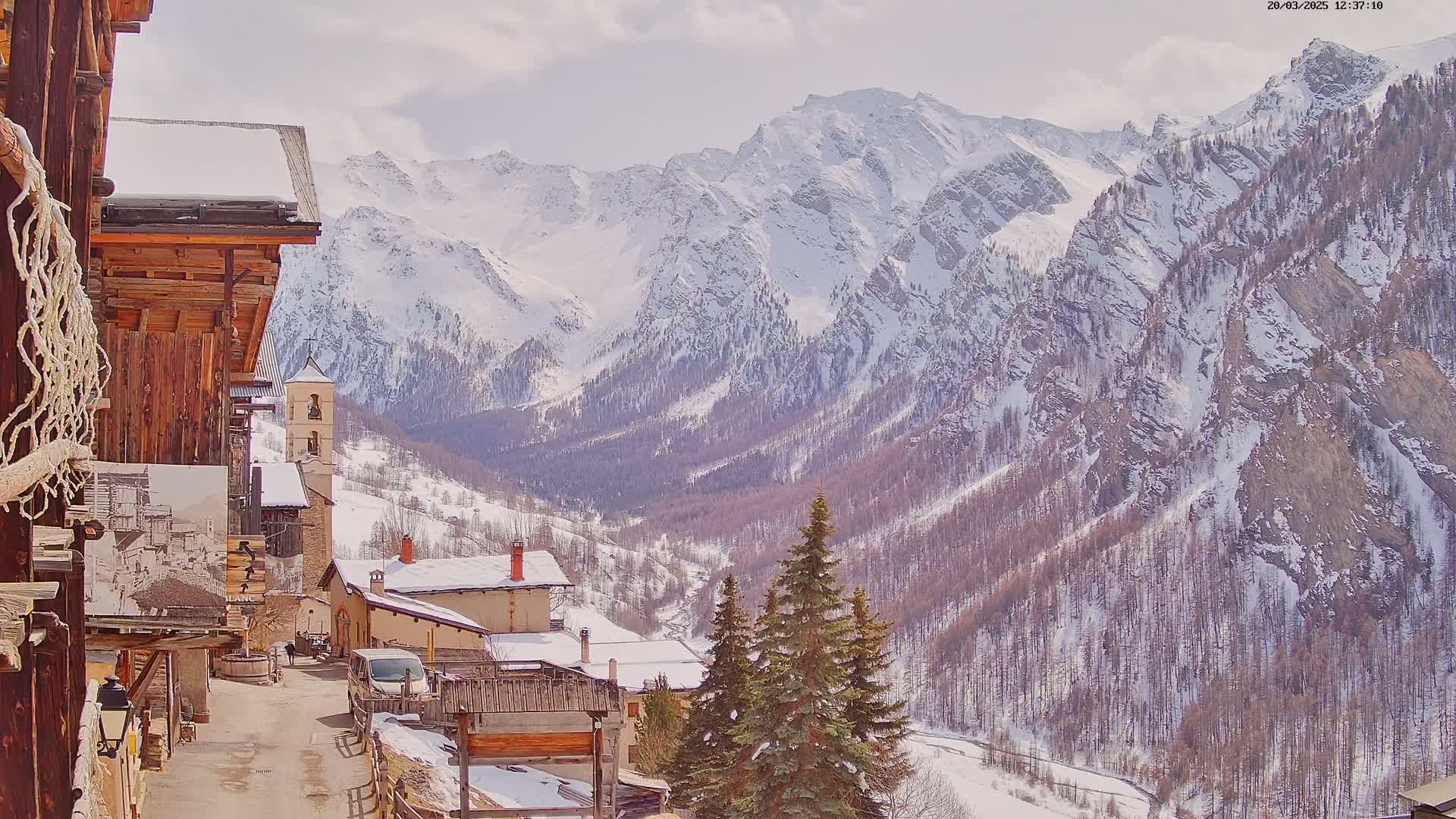 A snow-covered mountain valley is seen from a village of wooden buildings, under partly cloudy skies.