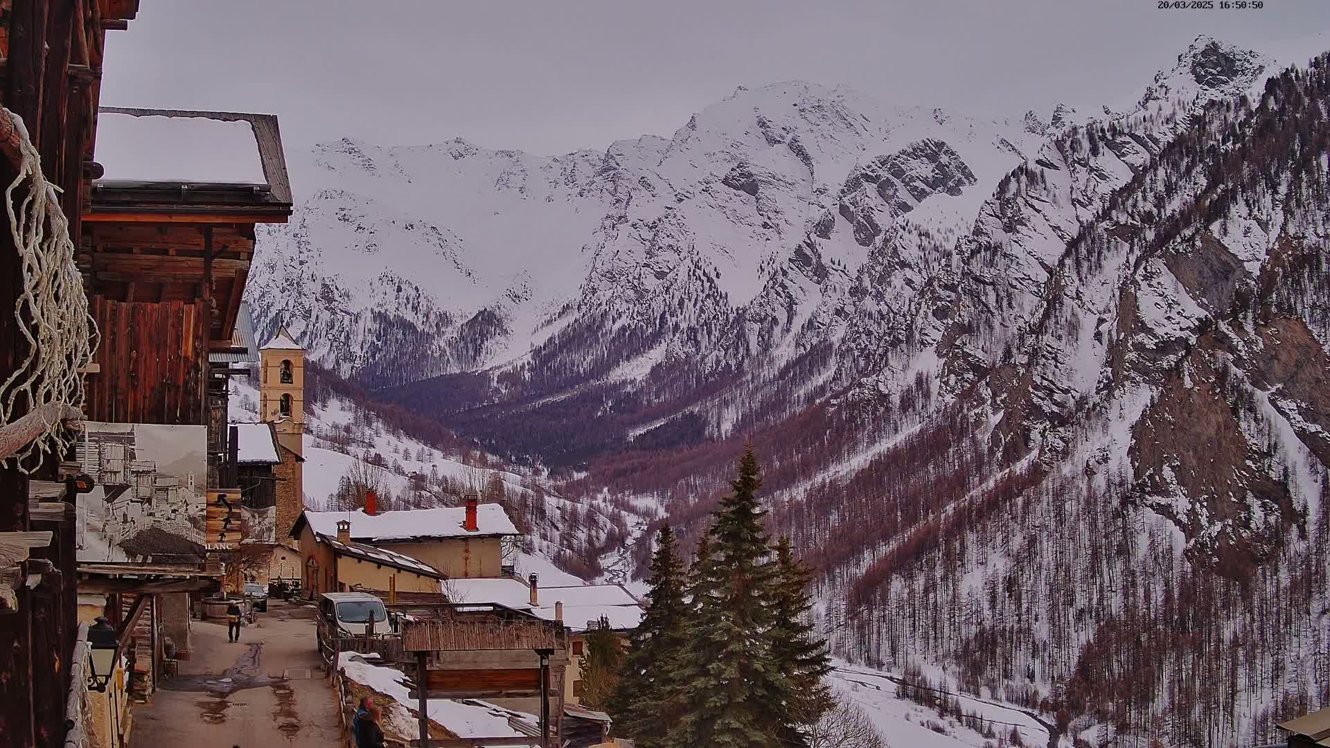 A snow-covered mountain village with a bell tower is nestled in a valley surrounded by snow-capped mountains under an overcast sky.