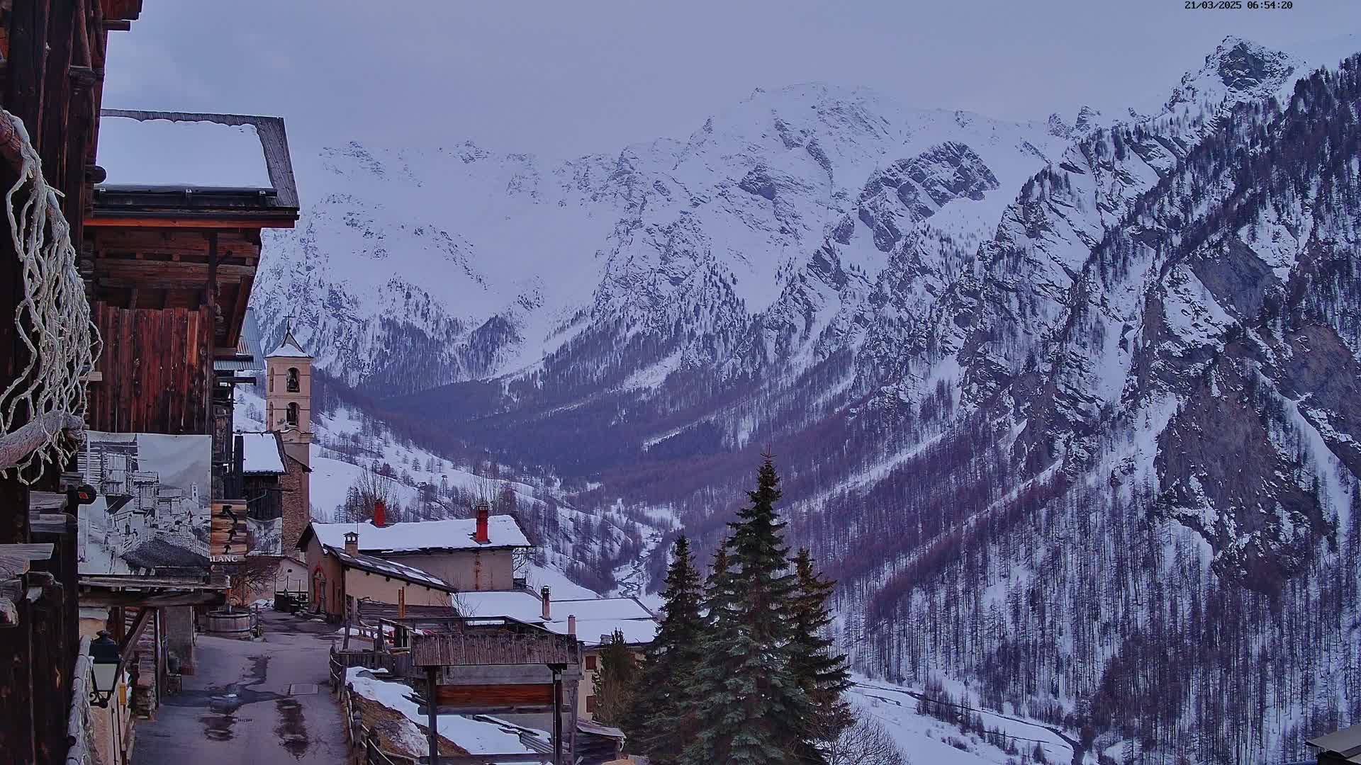 A snow-covered mountain valley is viewed from a village street, where several buildings with snow-covered roofs are visible under an overcast sky.