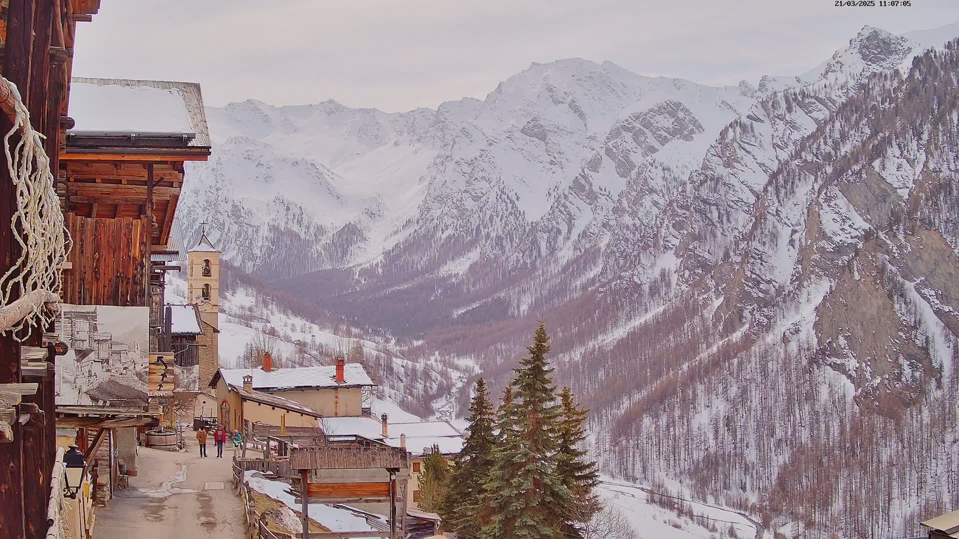 A snow-covered mountain valley is viewed from a village with rustic wooden buildings, a church steeple, and evergreen trees.