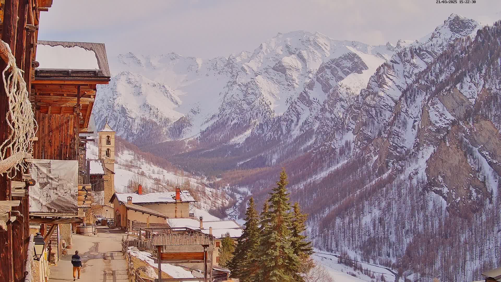 A snow-covered mountain valley is viewed from a village of rustic wooden buildings, under a mostly sunny sky.