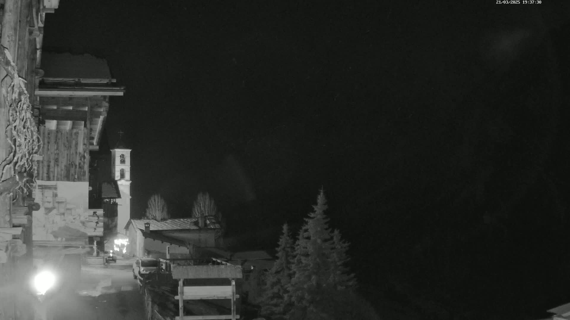 A nighttime, snow-dusted village scene shows a church steeple, buildings, and evergreens under a dark, possibly snowing sky.