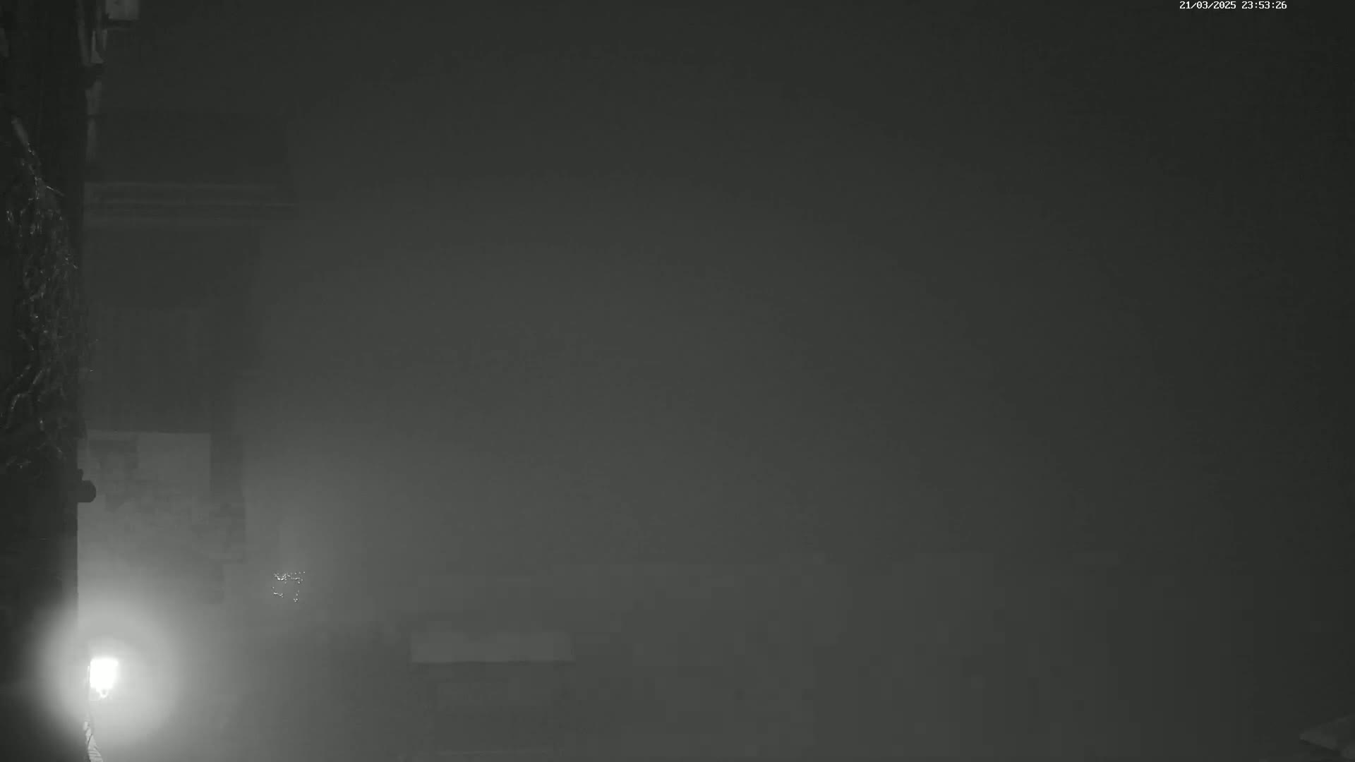 A dimly lit, foggy outdoor scene shows a building wall to the left with a light source near the bottom, and a hazy, indistinct space extending to the right.
