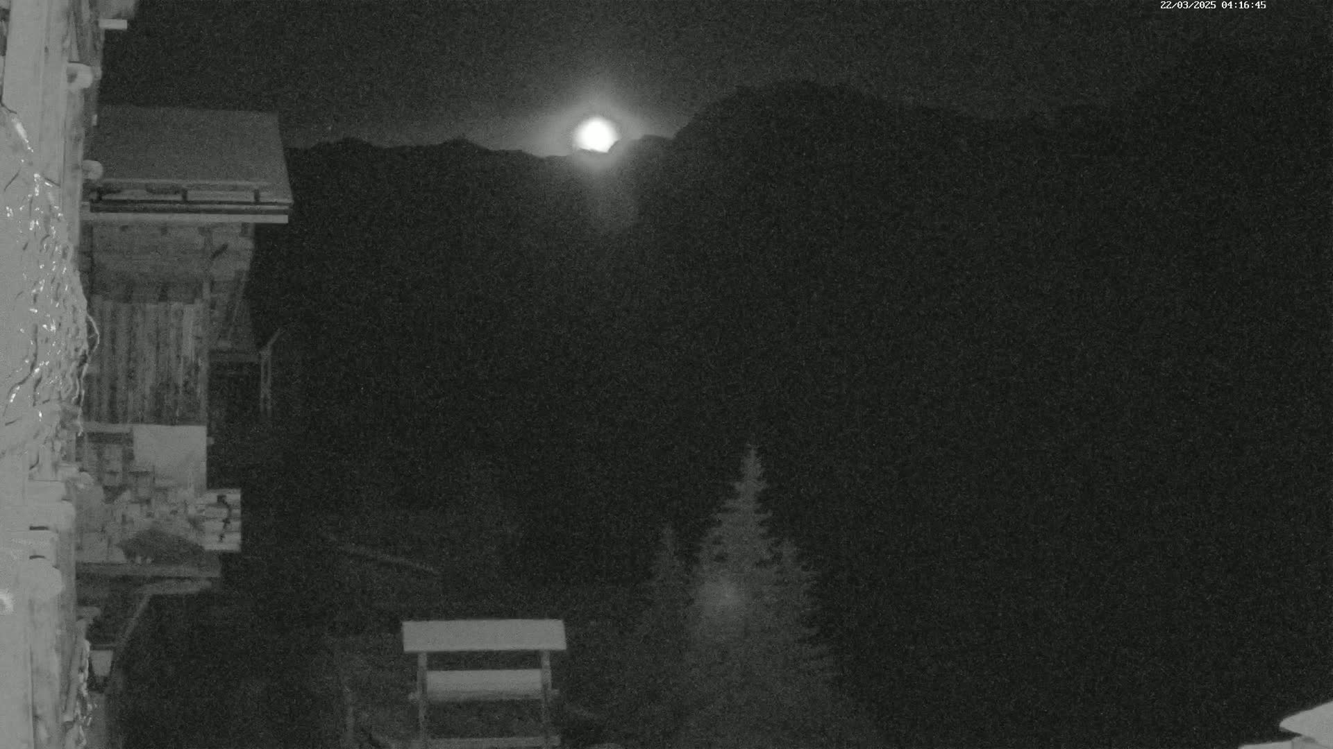 A bright moon illuminates a snow-covered wooden building and dark mountains at night.