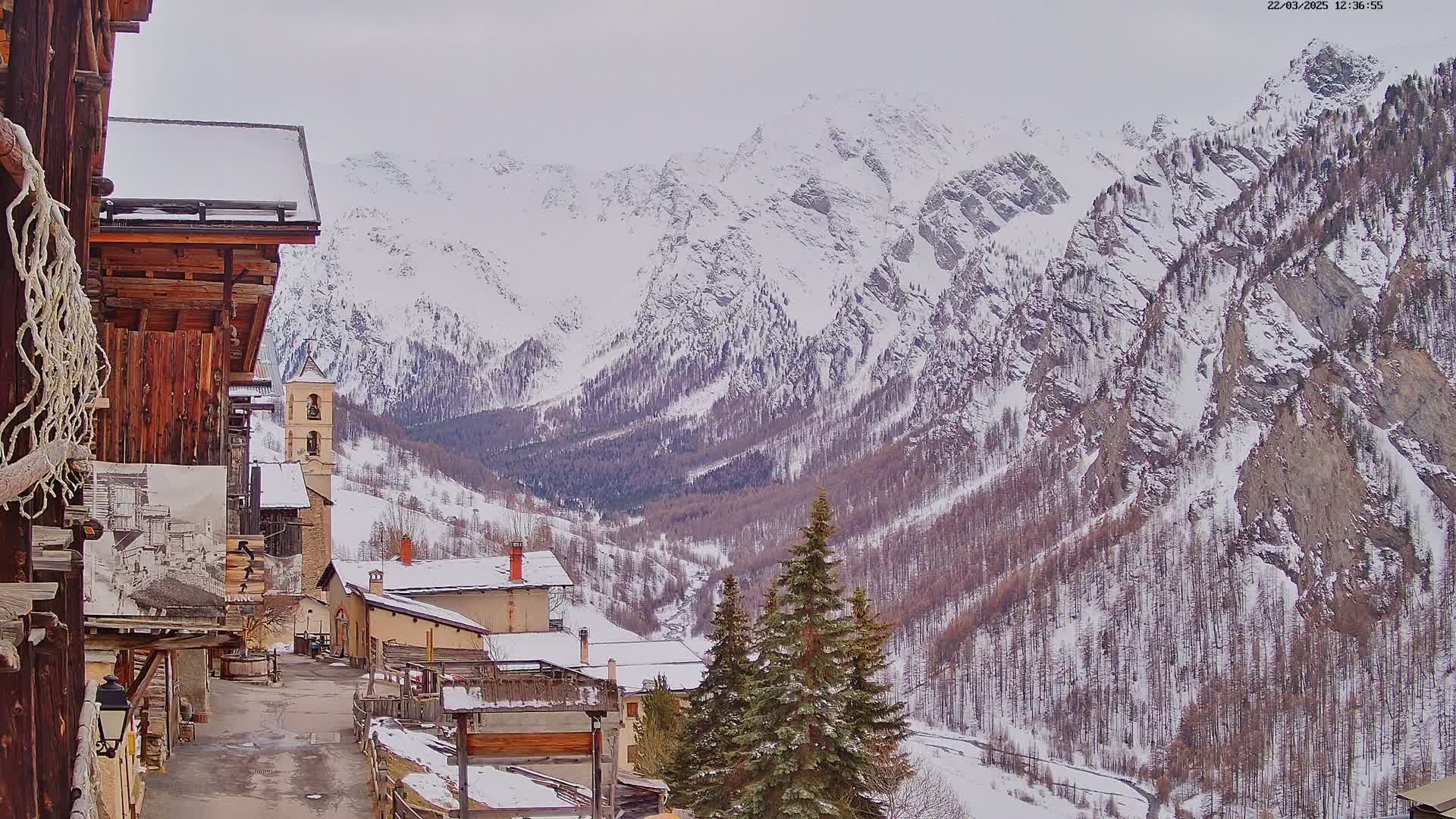 A snow-covered village nestled in a valley between towering, snow-capped mountains under an overcast sky.
