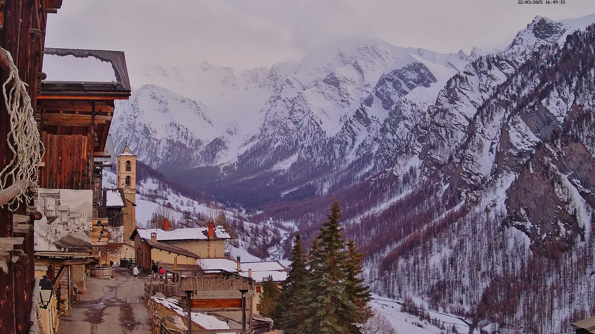 A snow-covered mountain valley is viewed from a village of rustic wooden buildings, with overcast skies.