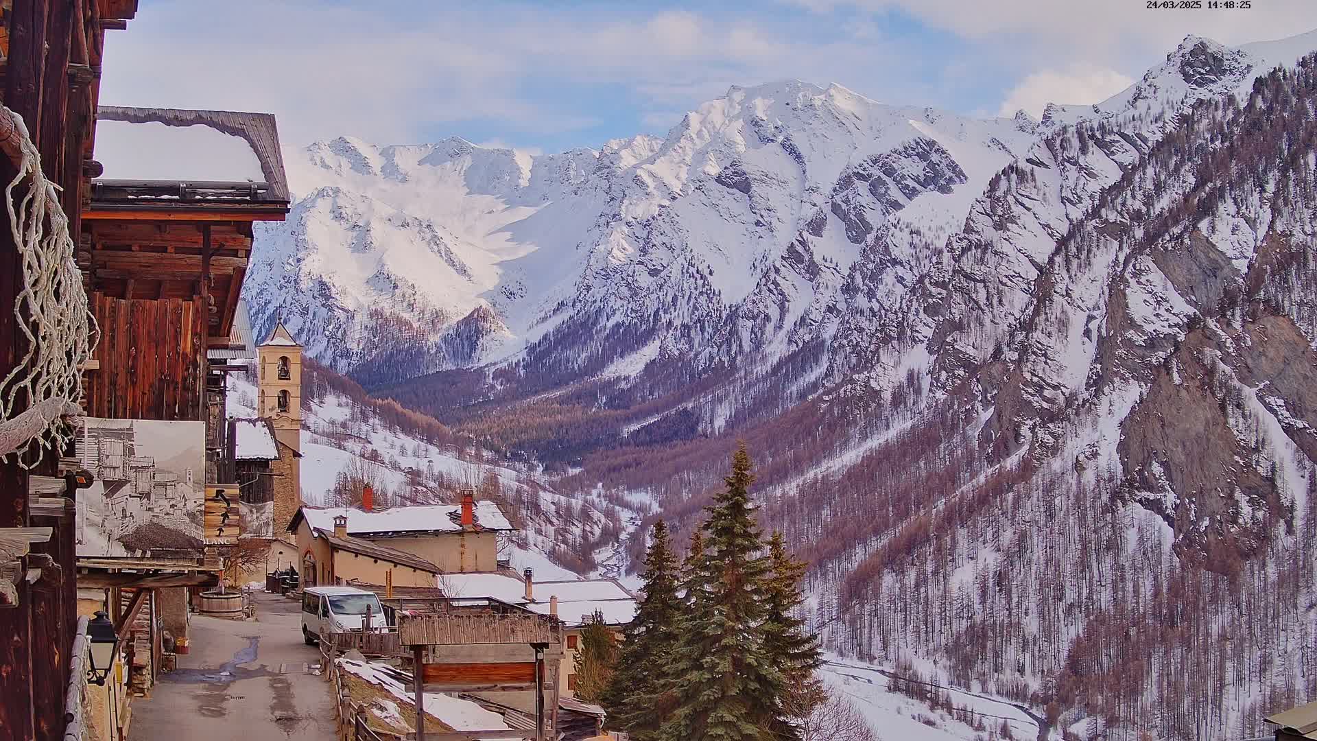 A snow-covered mountain valley village is seen from a roadside vantage point, with a partly sunny sky.