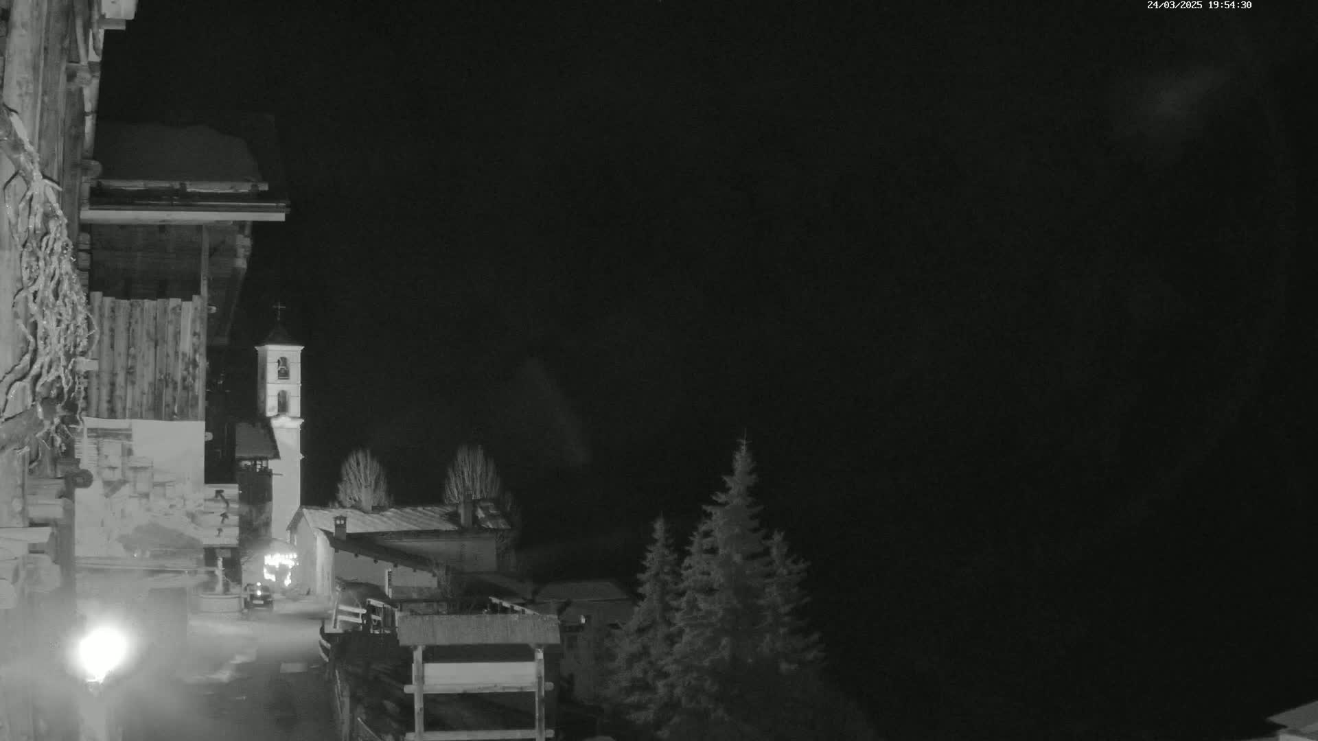 A nighttime, grayscale view of a snow-dusted village with a church steeple, some evergreen trees, and a building with a snow-covered roof in the foreground, under a dark, possibly cloudy sky.