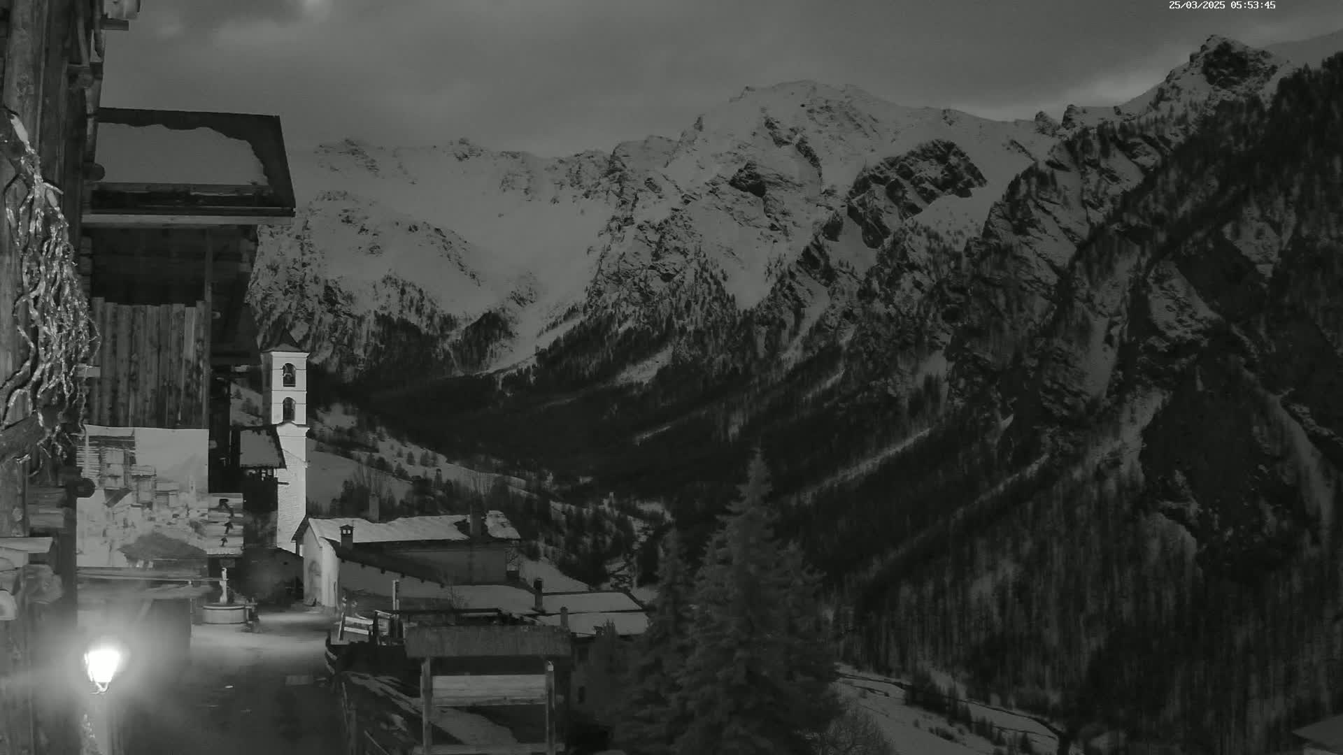A snow-covered mountain range overlooks a small village at night, illuminated by a single streetlamp.