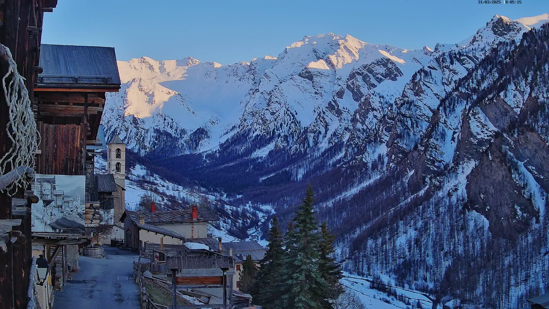 A snow-covered mountain range at sunset overlooks a small village of old stone buildings nestled in a valley.