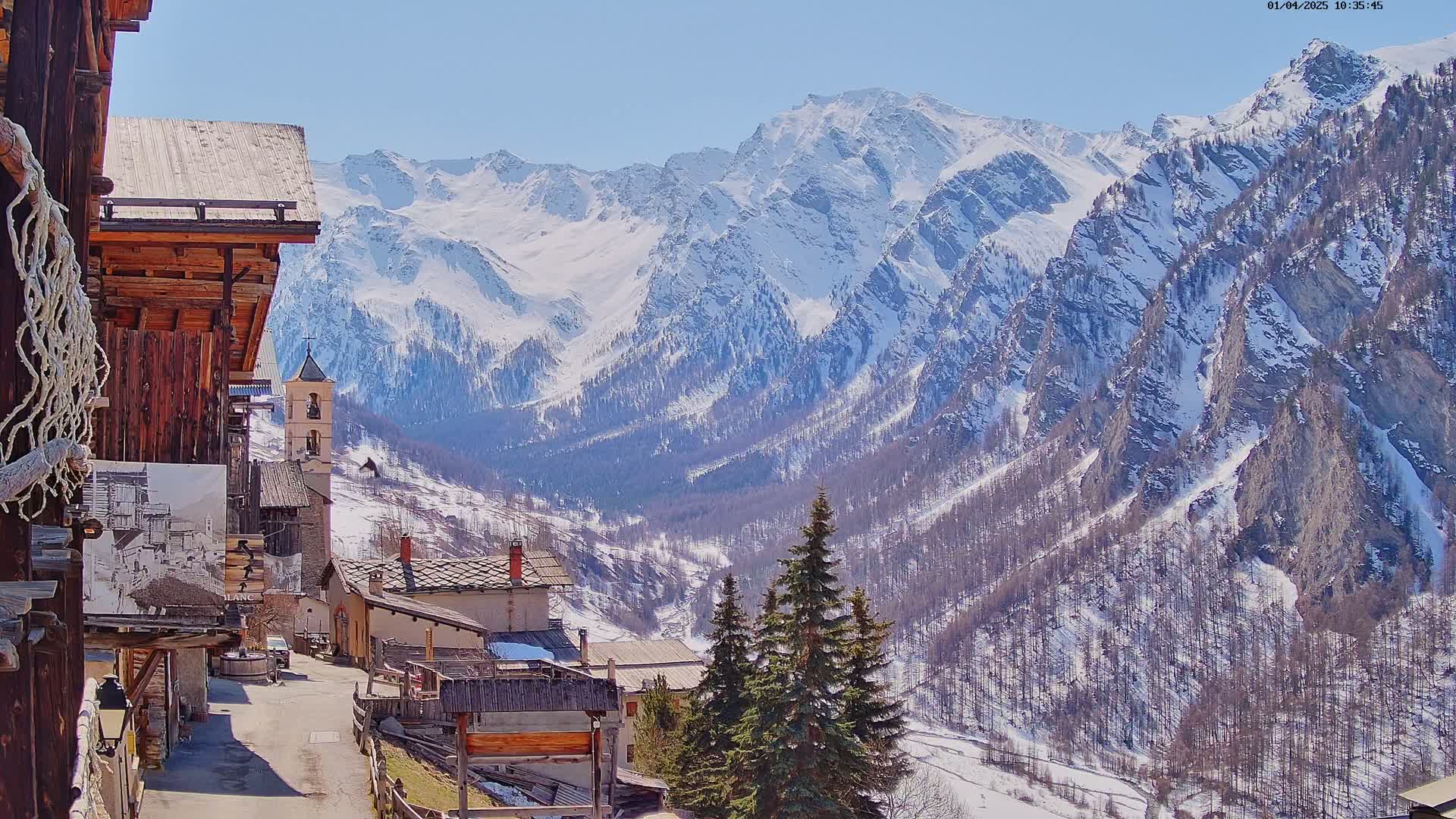 A snow-capped mountain range overlooks a small village nestled in a valley on a sunny day.