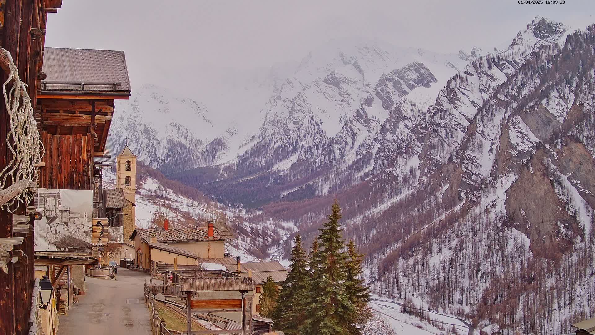 A snow-covered mountain valley is viewed from a village of rustic wooden and stone buildings on an overcast day.