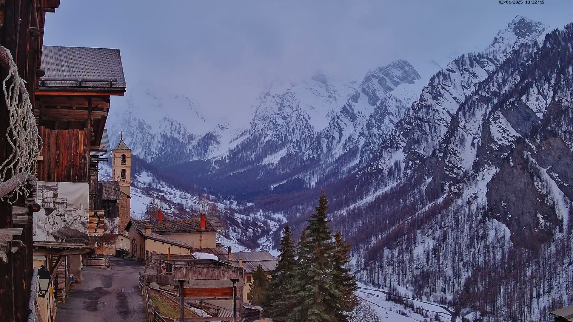 A snow-covered mountain valley is viewed from a village of old stone buildings, under an overcast sky.