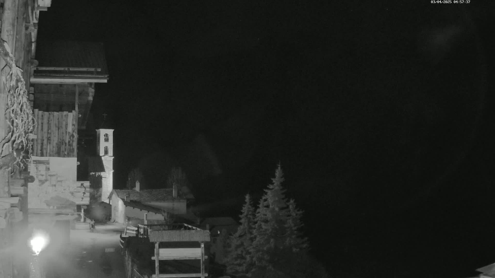 A nighttime, black-and-white view of a village scene shows a church steeple and several buildings, with dark evergreens in the background under a clear, dark sky.