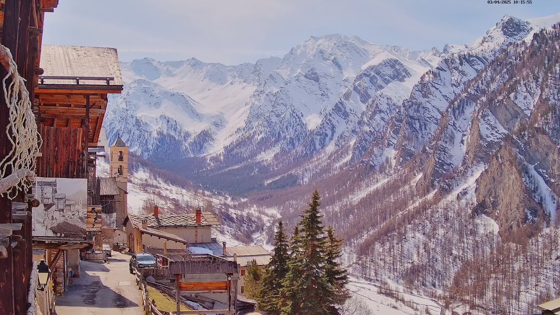 A snow-covered mountain range overlooks a small village with traditional wooden buildings under a bright, sunny sky.