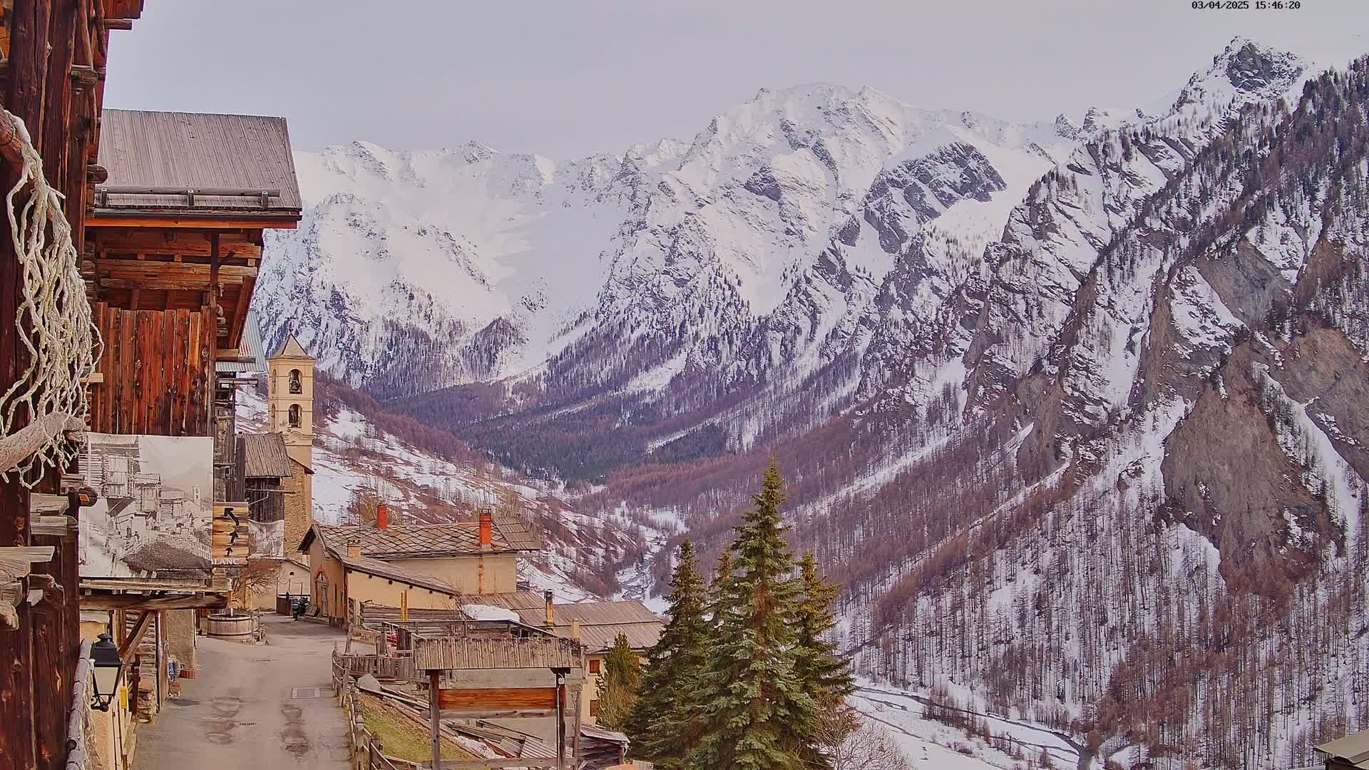A quaint village nestled in a valley, with snow-covered mountains rising in the background under a cloudy sky.