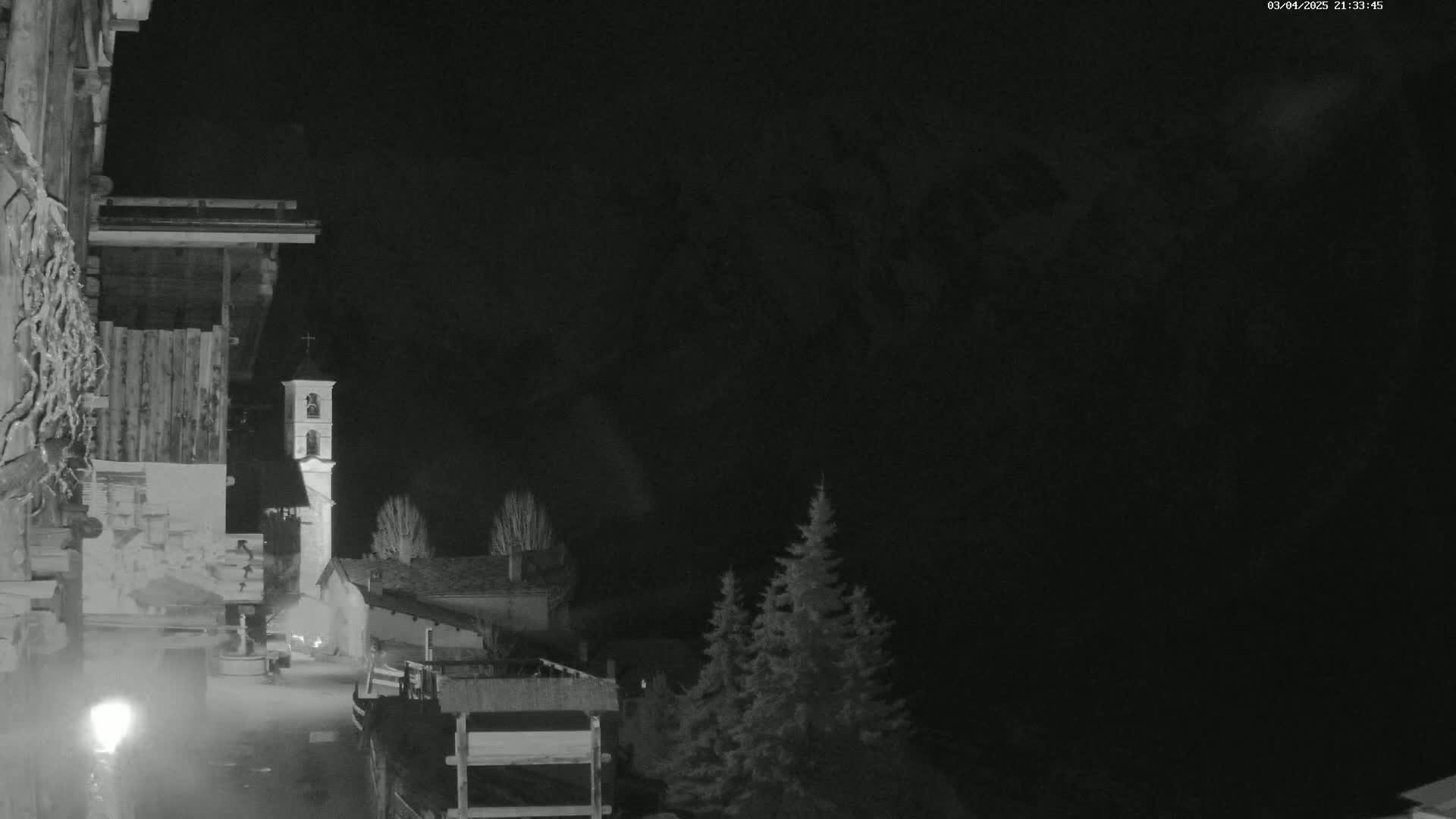 A nighttime, black-and-white view of a village with a church steeple, partially obscured by darkness and a few evergreens.