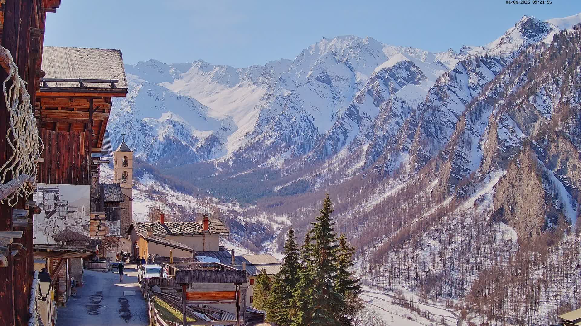 A snow-capped mountain range is visible behind a small village nestled in a valley on a sunny day.