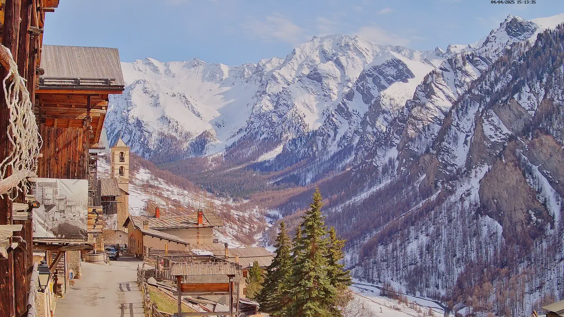 A snow-capped mountain range overlooks a quaint village nestled in a valley on a sunny day.