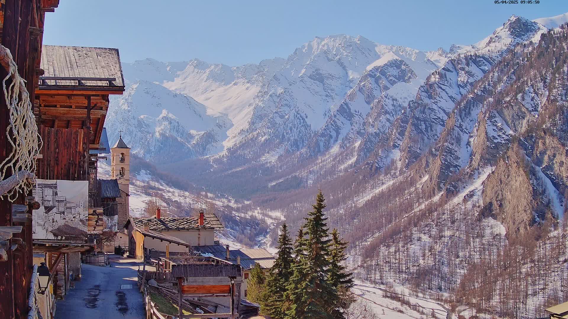 A snow-covered mountain range rises behind a quaint village of rustic wooden buildings on a sunny day.
