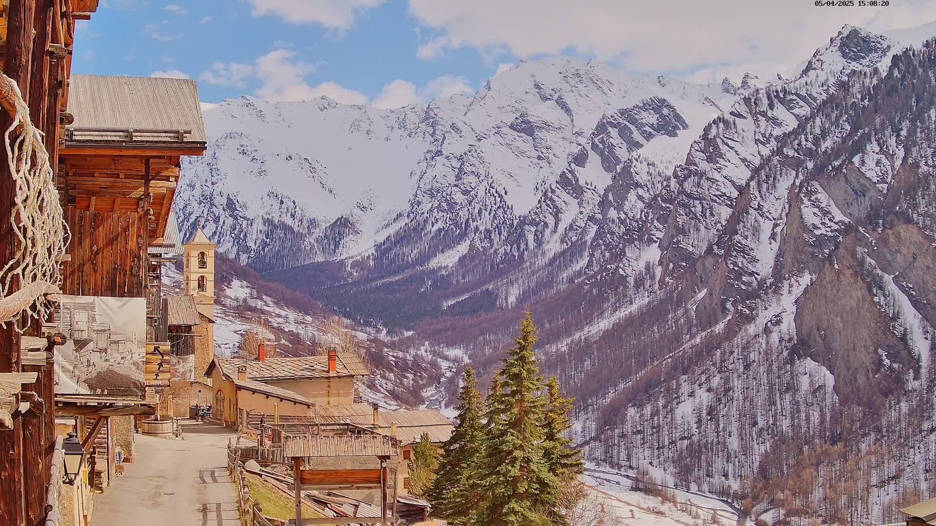 A quaint village nestled in a valley, surrounded by snow-capped mountains under a partly cloudy sky.