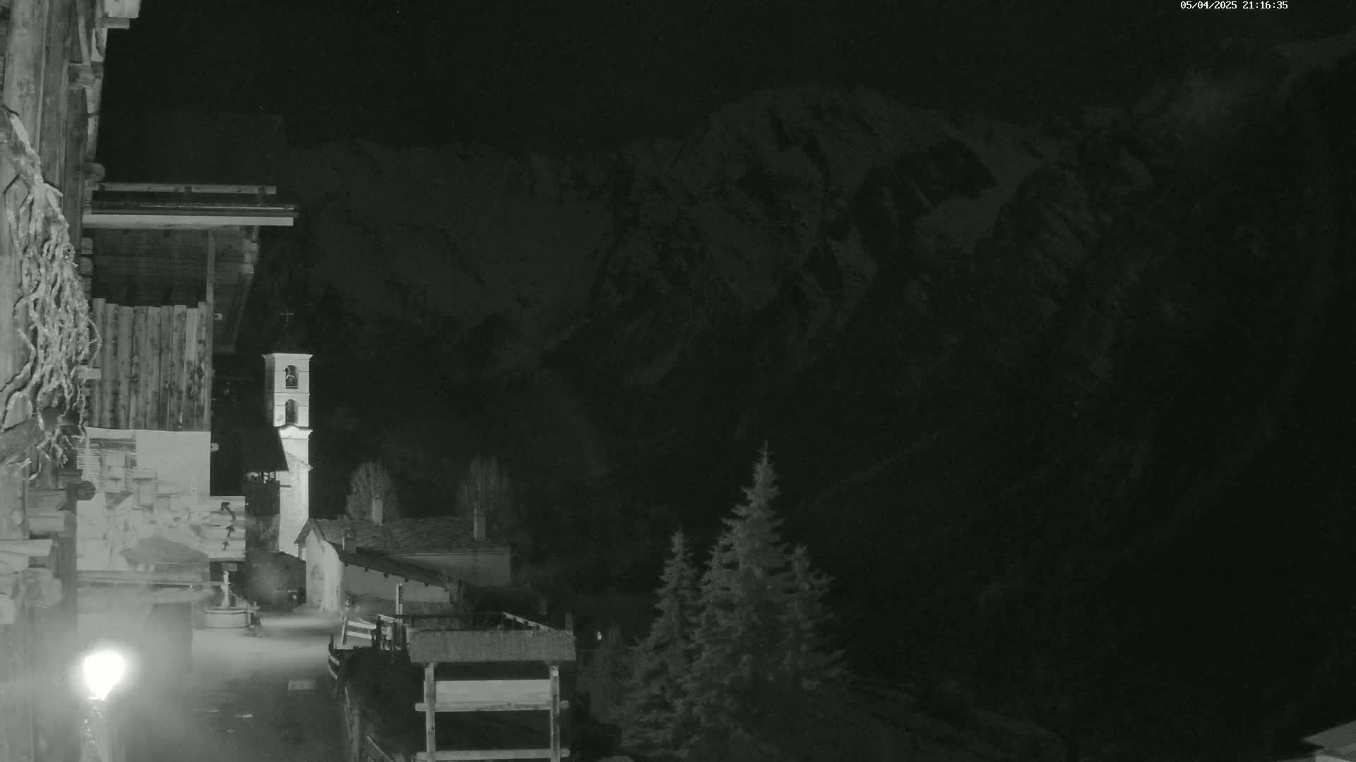 A nighttime view of a village nestled at the base of snow-capped mountains, illuminated by a single lamppost.
