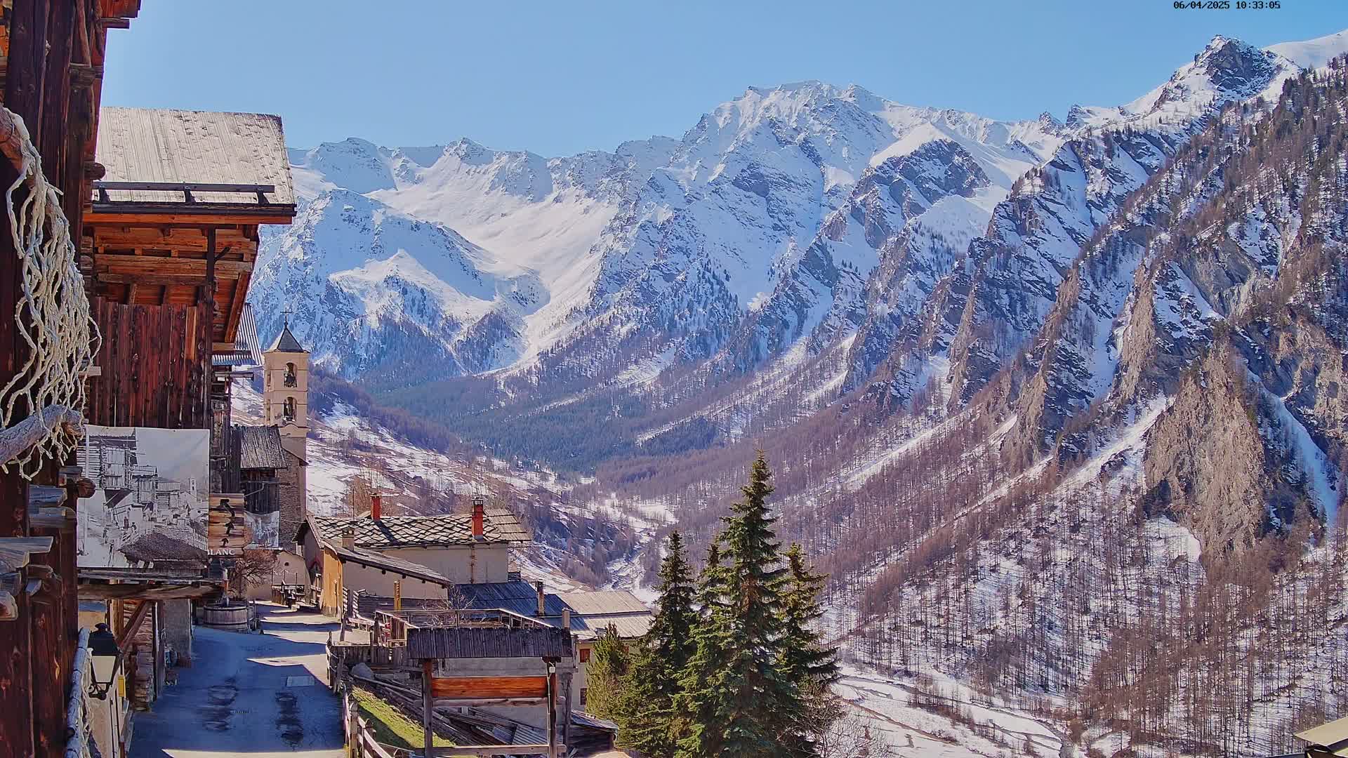 A snow-capped mountain range looms over a small village nestled in a valley on a sunny day.