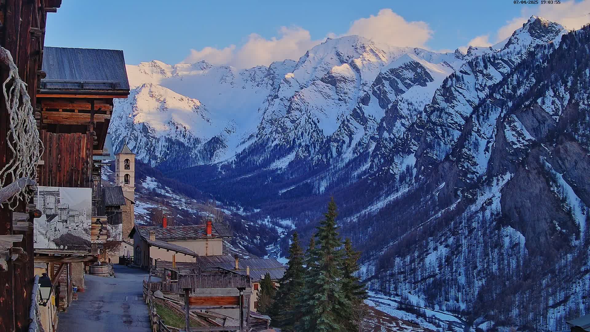 A snow-capped mountain range overlooks a small village of old stone buildings under a mostly clear, blue sky.