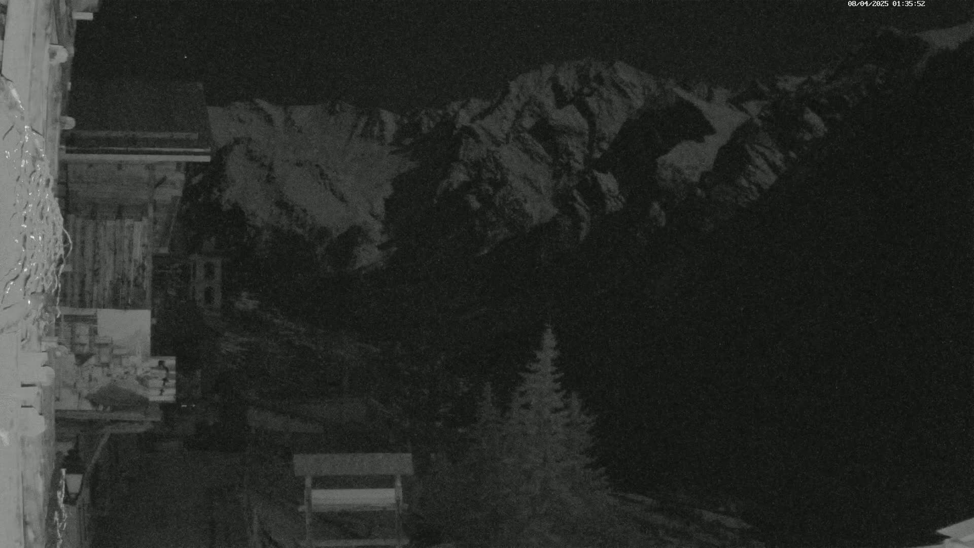 A nighttime view shows snow-covered mountains in the distance, with a wooden structure and evergreen trees in the foreground under a dark sky.