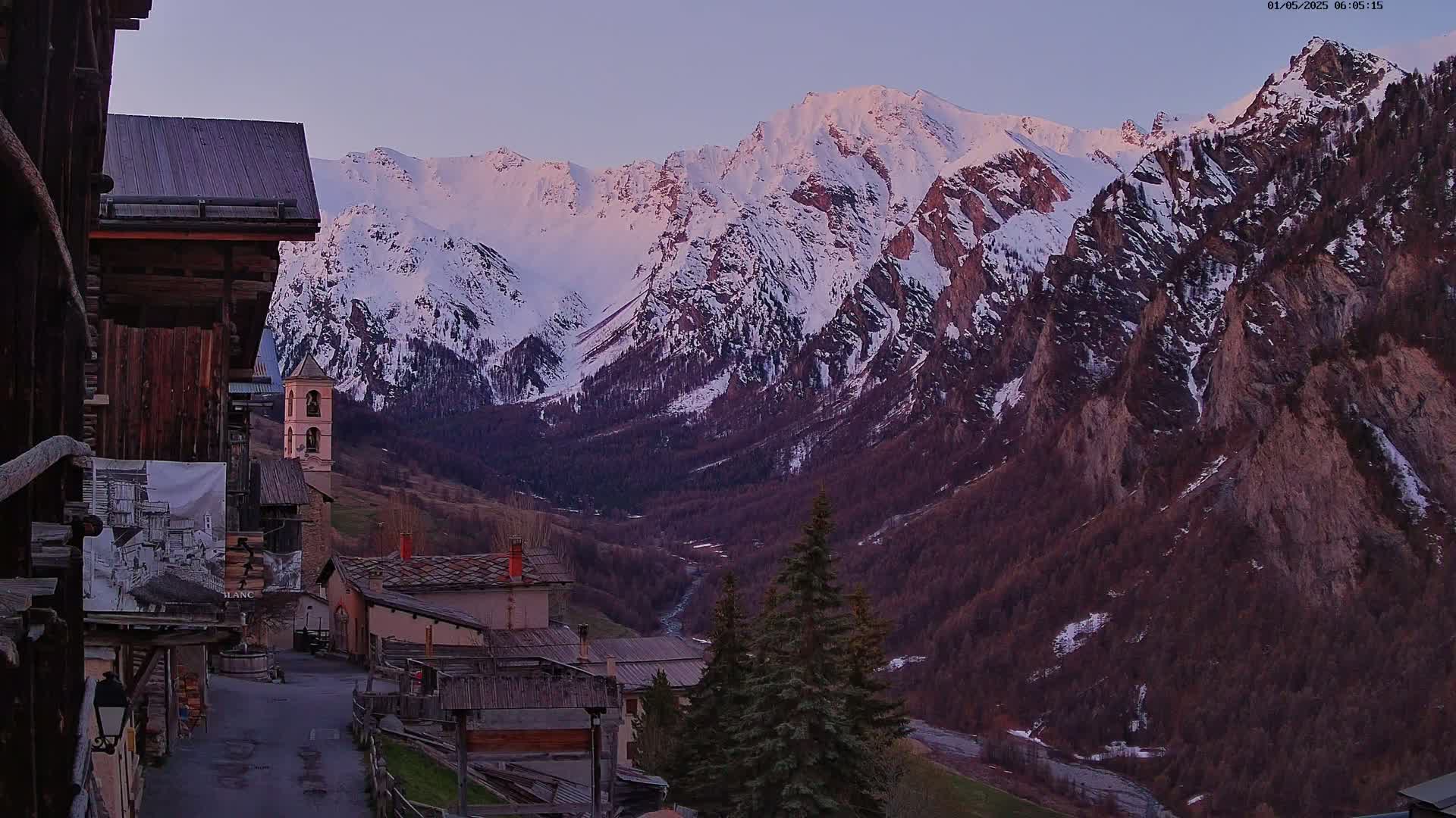 A mountain village with rustic wooden buildings nestles in a valley at dawn, overlooked by snow-capped mountains under a clear, pale sky.