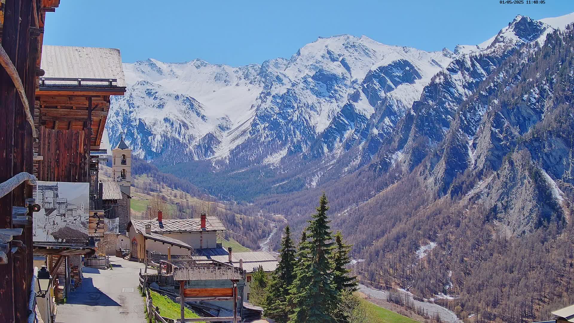 A quaint mountain village nestled in a valley, with snow-capped mountains under a clear blue sky.