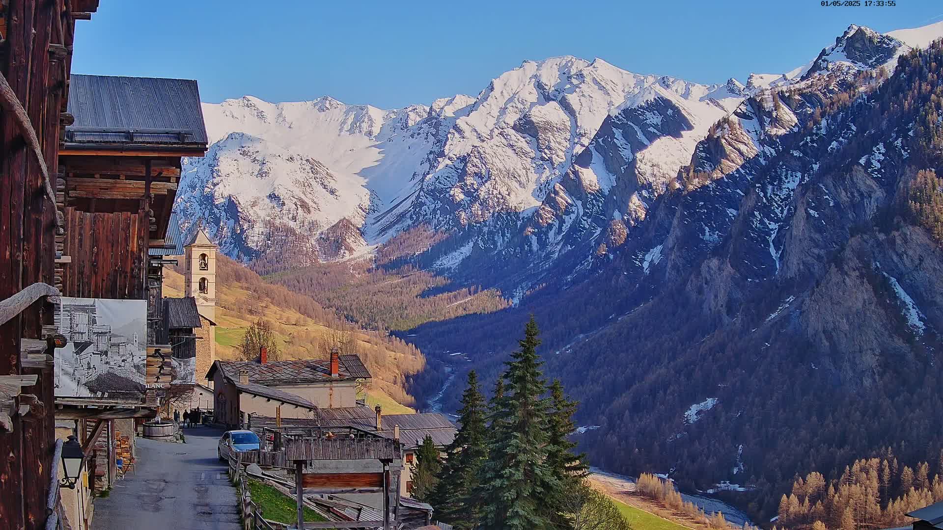 A quaint mountain village with wooden buildings nestles in a valley, overlooked by snow-capped mountains under a clear blue sky.